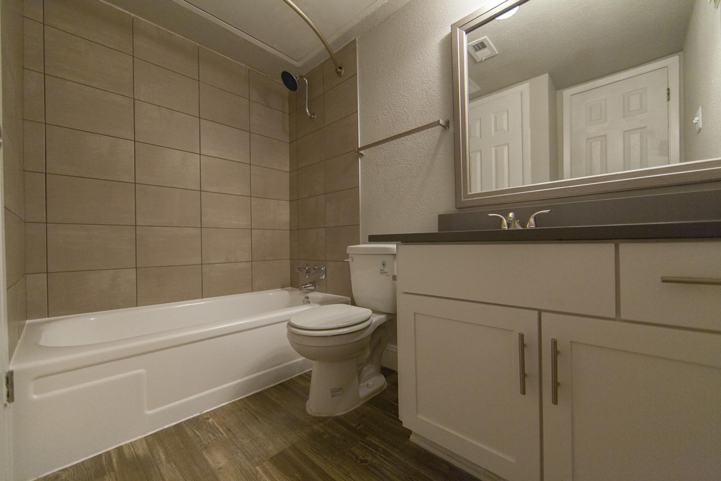 A modern bathroom featuring a bathtub with shower, a white toilet, and a sleek vanity with a mirror. The walls are tiled with beige tiles, and the floor has a wood-like finish. The space is well-lit, creating a clean and inviting atmosphere.