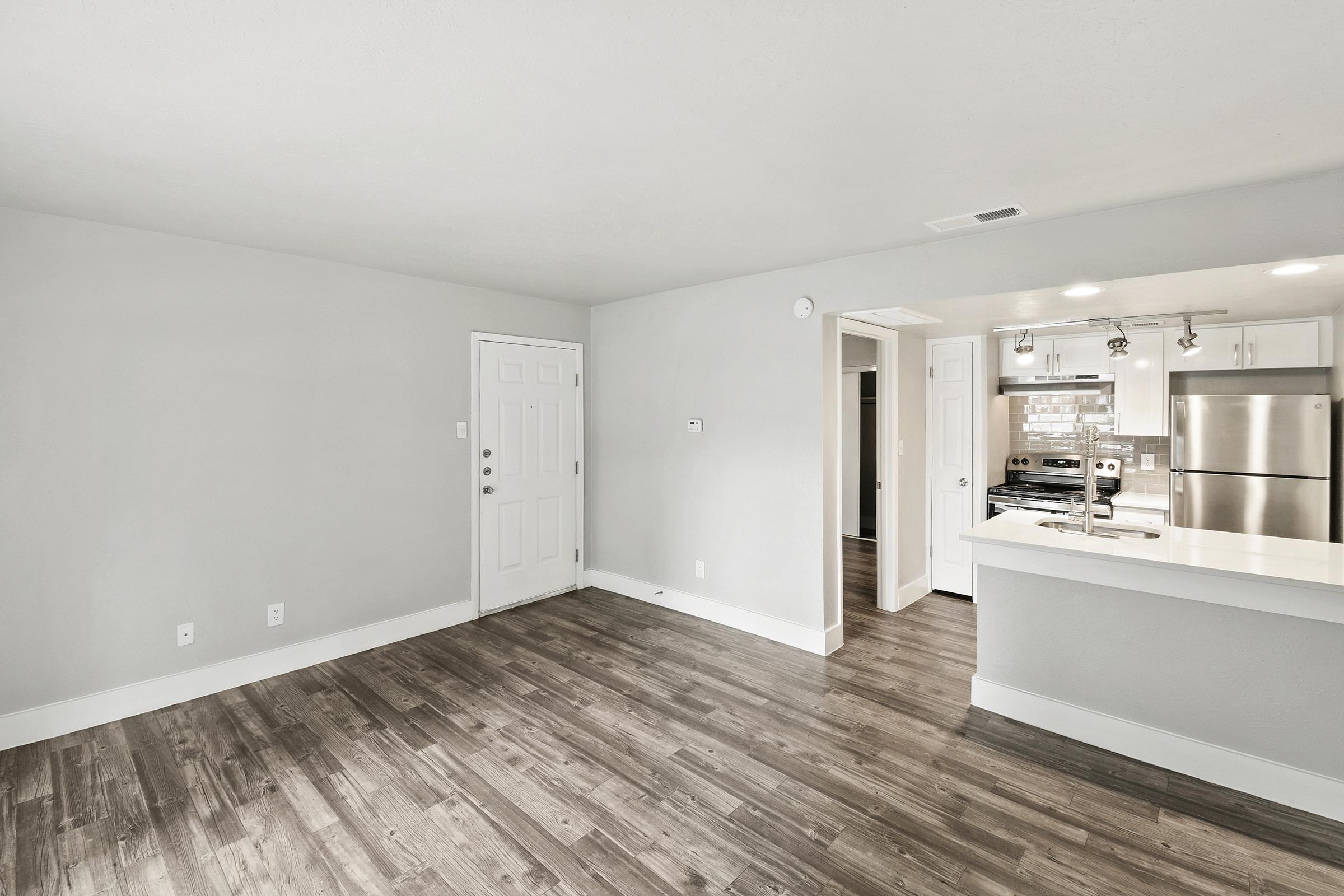 A spacious, modern living area with light gray walls and hardwood flooring. The room features a front door, a kitchenette with stainless steel appliances, and a breakfast bar. Natural light fills the space, creating a bright and inviting atmosphere.