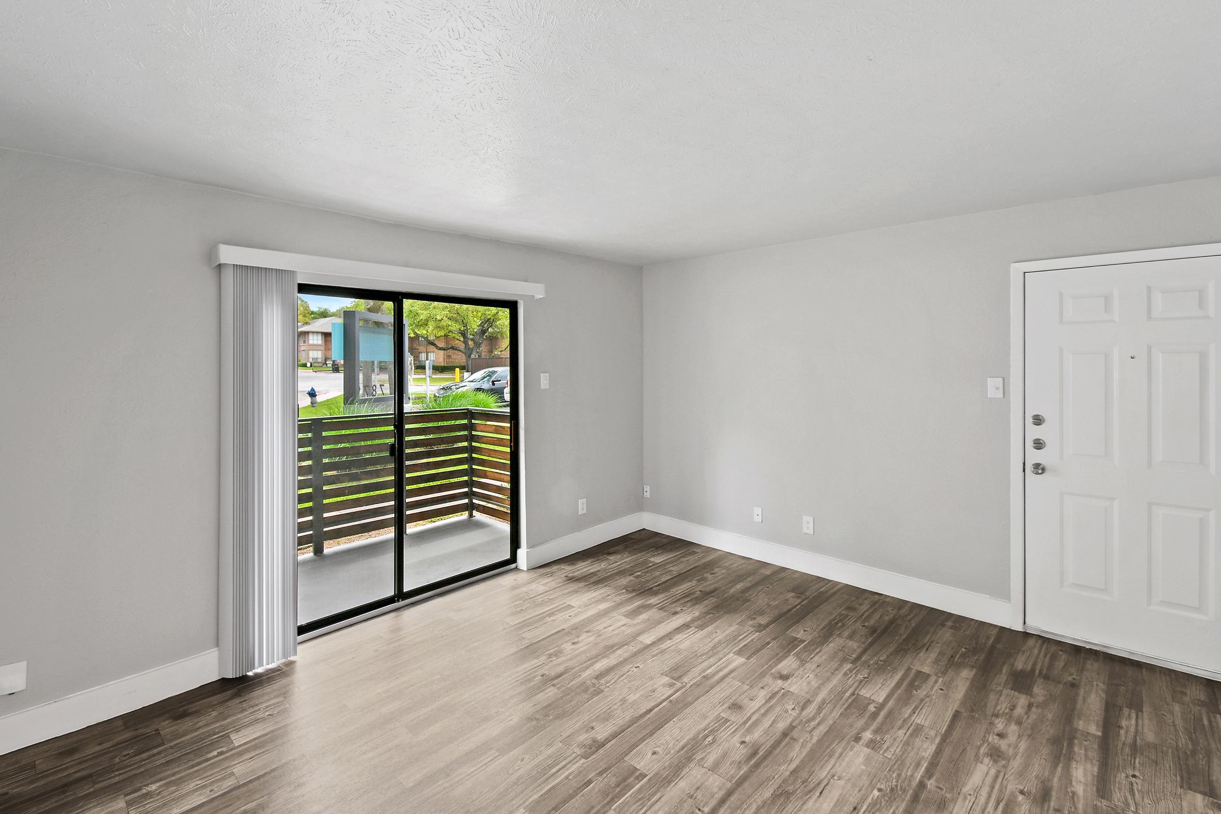 A light-filled interior of a living room with a sliding glass door leading to a small balcony. The walls are painted in a soft gray, and the floor features wood-like laminate. The entrance door is white, and the space is empty, creating a fresh and welcoming atmosphere.