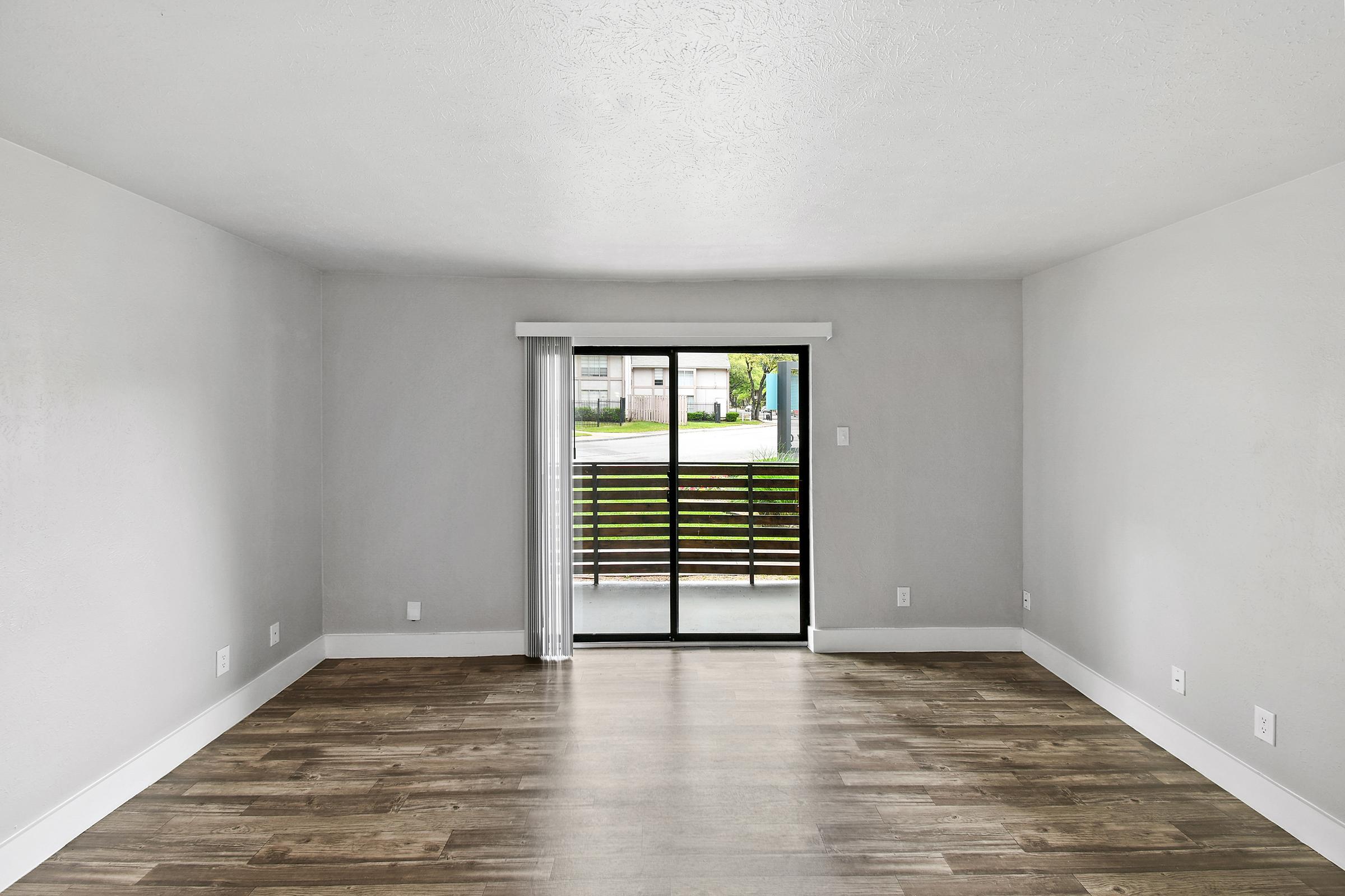 An empty living room with light gray walls and hardwood flooring. A large sliding glass door with vertical blinds opens to a small outdoor area. The natural light brightens the space, which has a minimalist feel. The room is spacious, with no furniture visible.