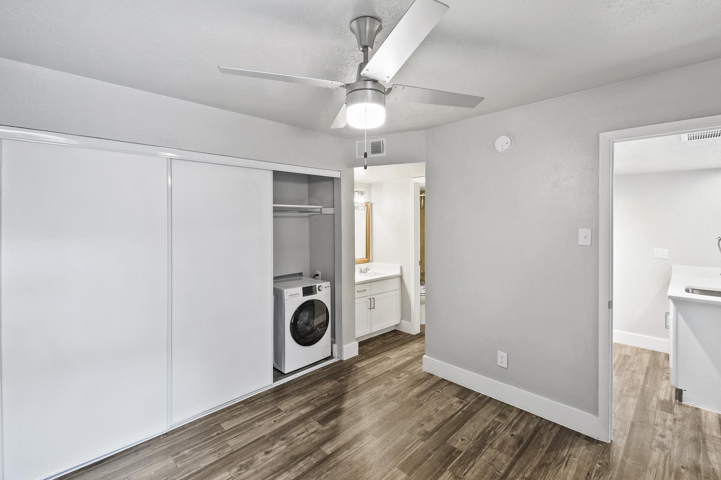 A spacious interior of a modern room featuring a washer and dryer in a closet, a bathroom doorway, and light-colored walls. The floor is a warm wood tone, and a ceiling fan is visible, enhancing the contemporary design. Natural light comes from an unseen window.