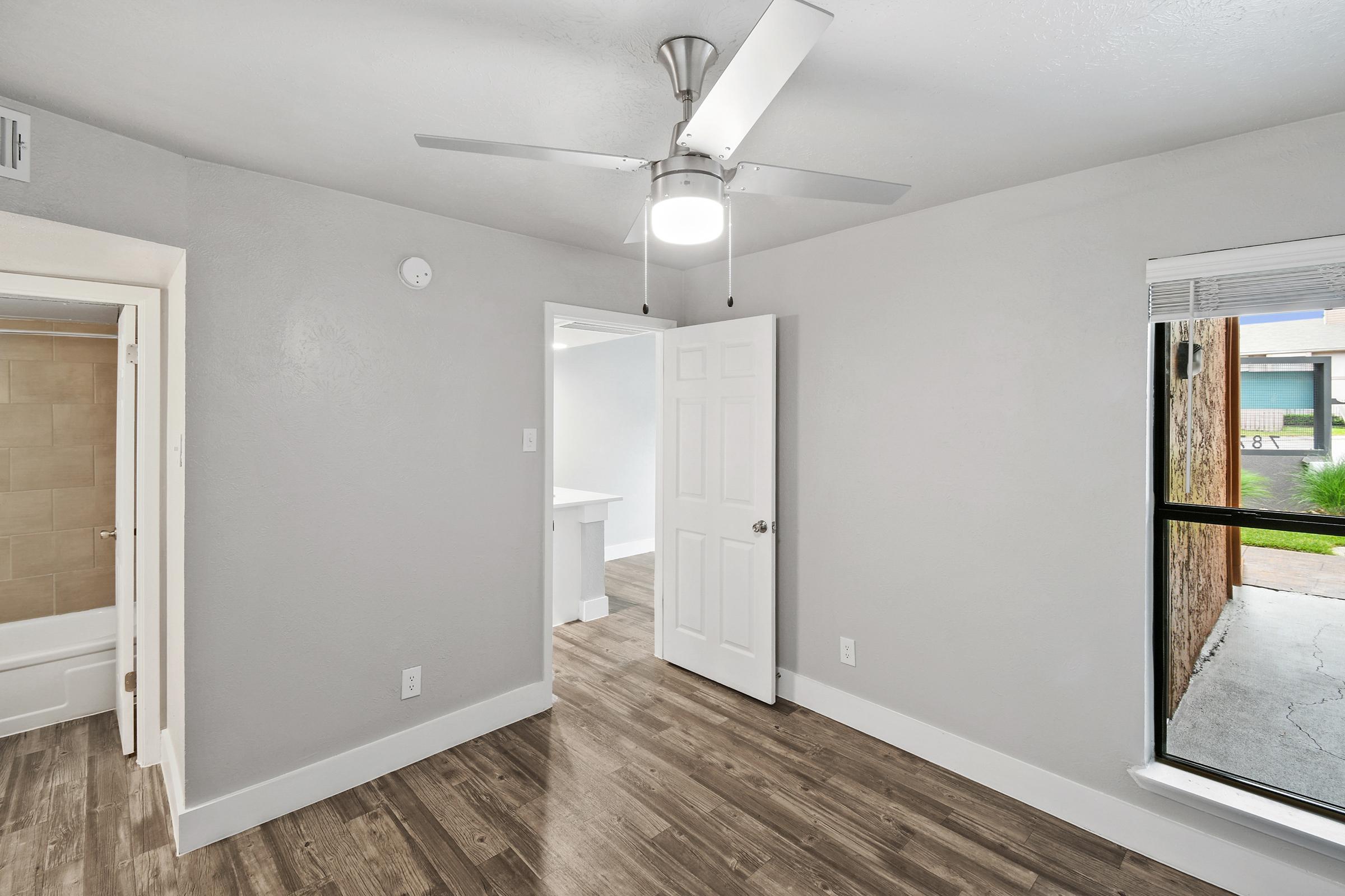 Interior of a light gray room with a ceiling fan and modern flooring. A doorway leads to another room, and a window lets in natural light, showing a view of the outside. The walls are bare, creating a minimalist aesthetic. A bathroom door is visible on the left.
