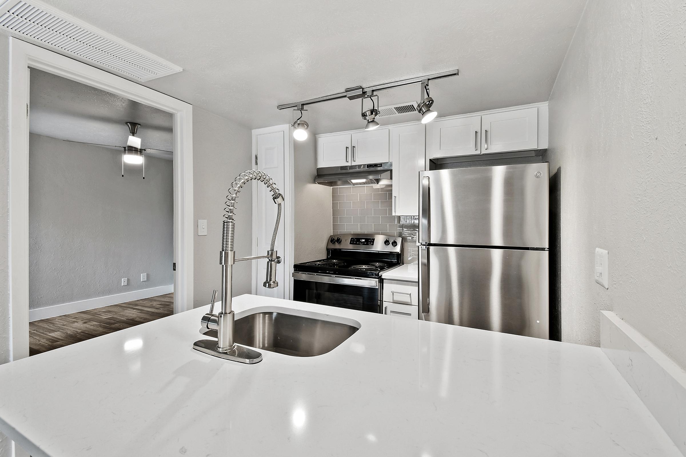 A modern kitchen featuring a sleek white countertop with a sink, stainless steel appliances including a refrigerator and an oven, and white cabinets. The space has track lighting and a neutral-colored wall, enhancing the contemporary design. In the background, a doorway leads to another room.