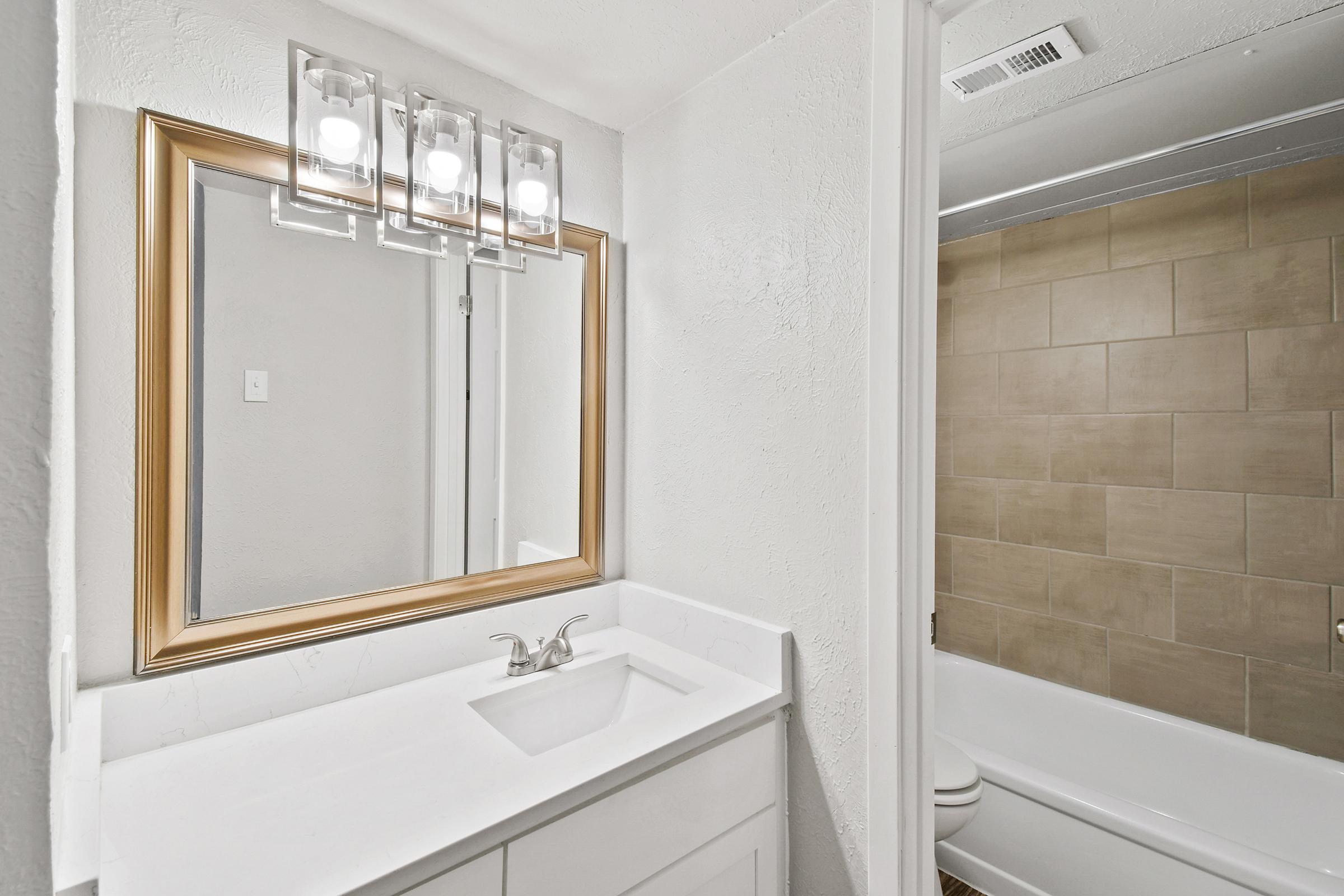 A modern bathroom featuring a clean white vanity with a rectangular sink, a large mirror framed in gold, and elegant light fixtures above. The space has light gray walls and beige tiled accents. To the right, a bathtub with a shower is visible, enhancing the overall bright and airy feel of the room.