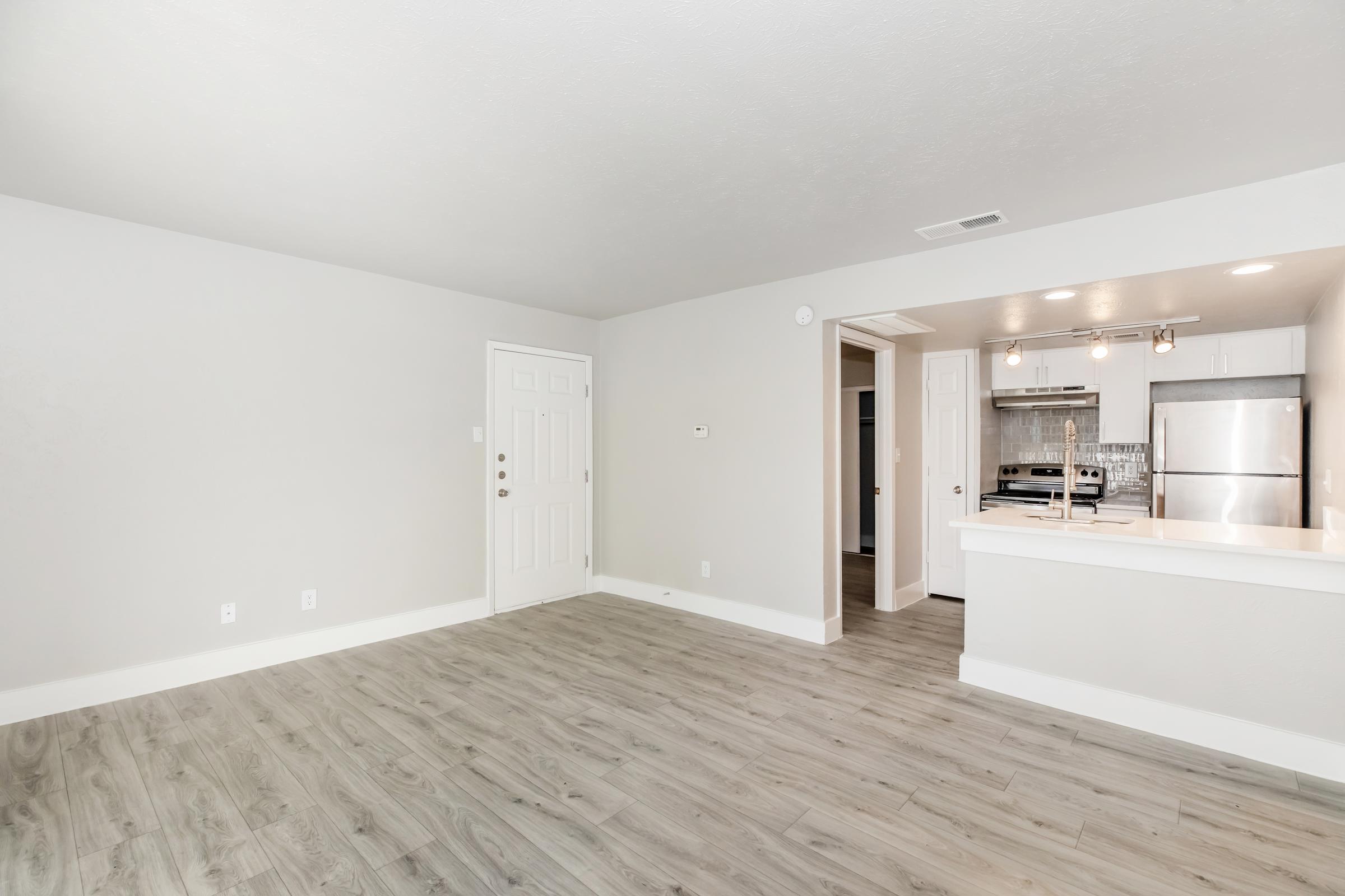 A spacious, empty living room with light gray walls and hardwood-style flooring. To the right, there's an open kitchen featuring stainless steel appliances and a bar counter. The entrance door is visible on the left, and there are modern light fixtures above the kitchen area. Natural light brightens the room.