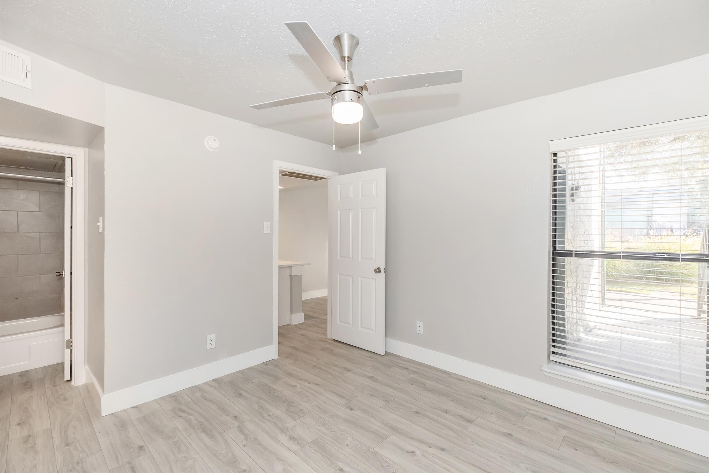 A well-lit room featuring light gray walls, a ceiling fan with a light fixture, and laminate flooring. There are two doors: one leading to a bathroom and another to another room. A large window with blinds allows natural light to enter the space.