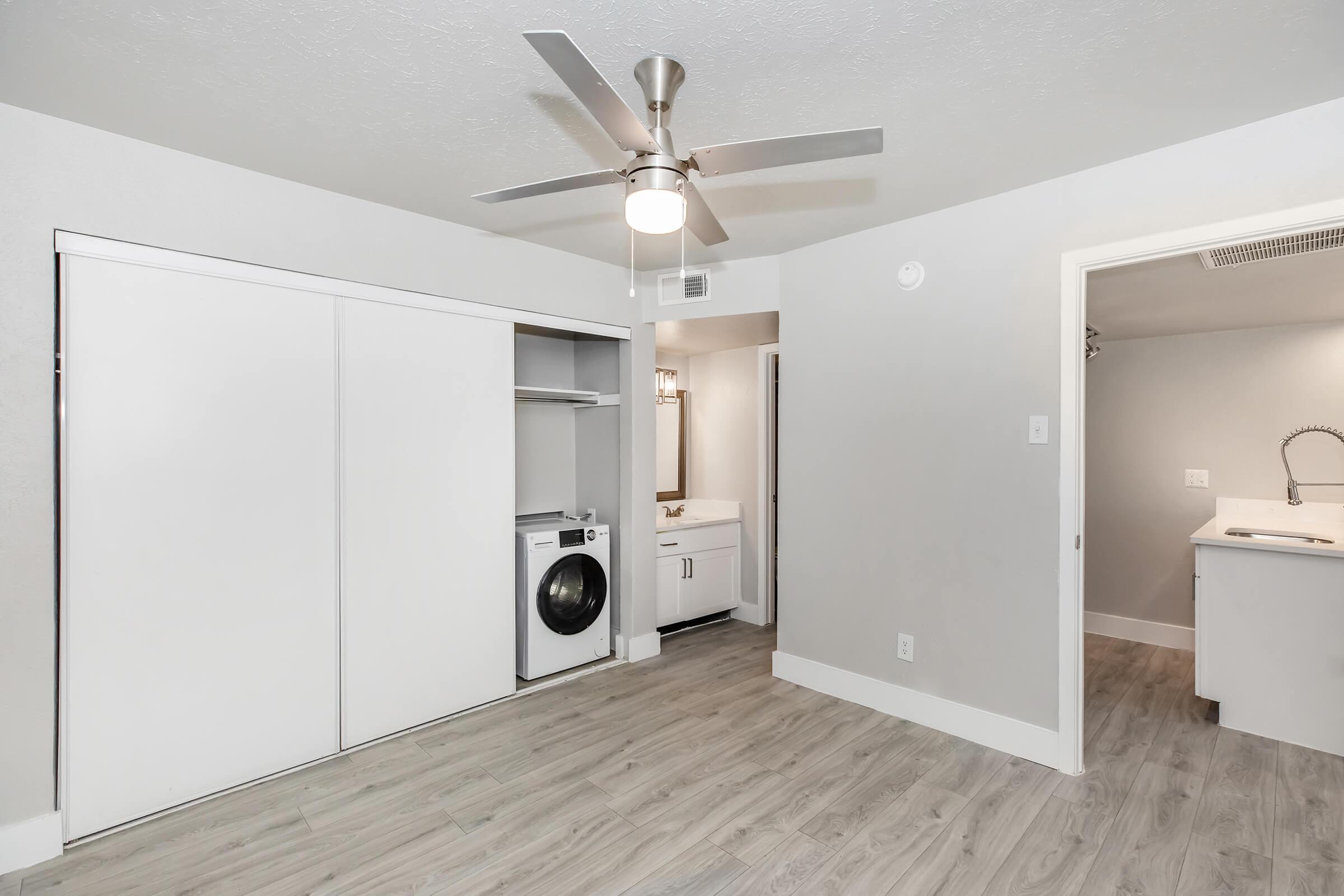 Interior view of a modern, light-colored room featuring a ceiling fan, laundry area with a washer, and a bathroom doorway. The walls are painted gray, and the flooring is a light wood look. There is a closet with sliding doors and a bright, clean aesthetic.