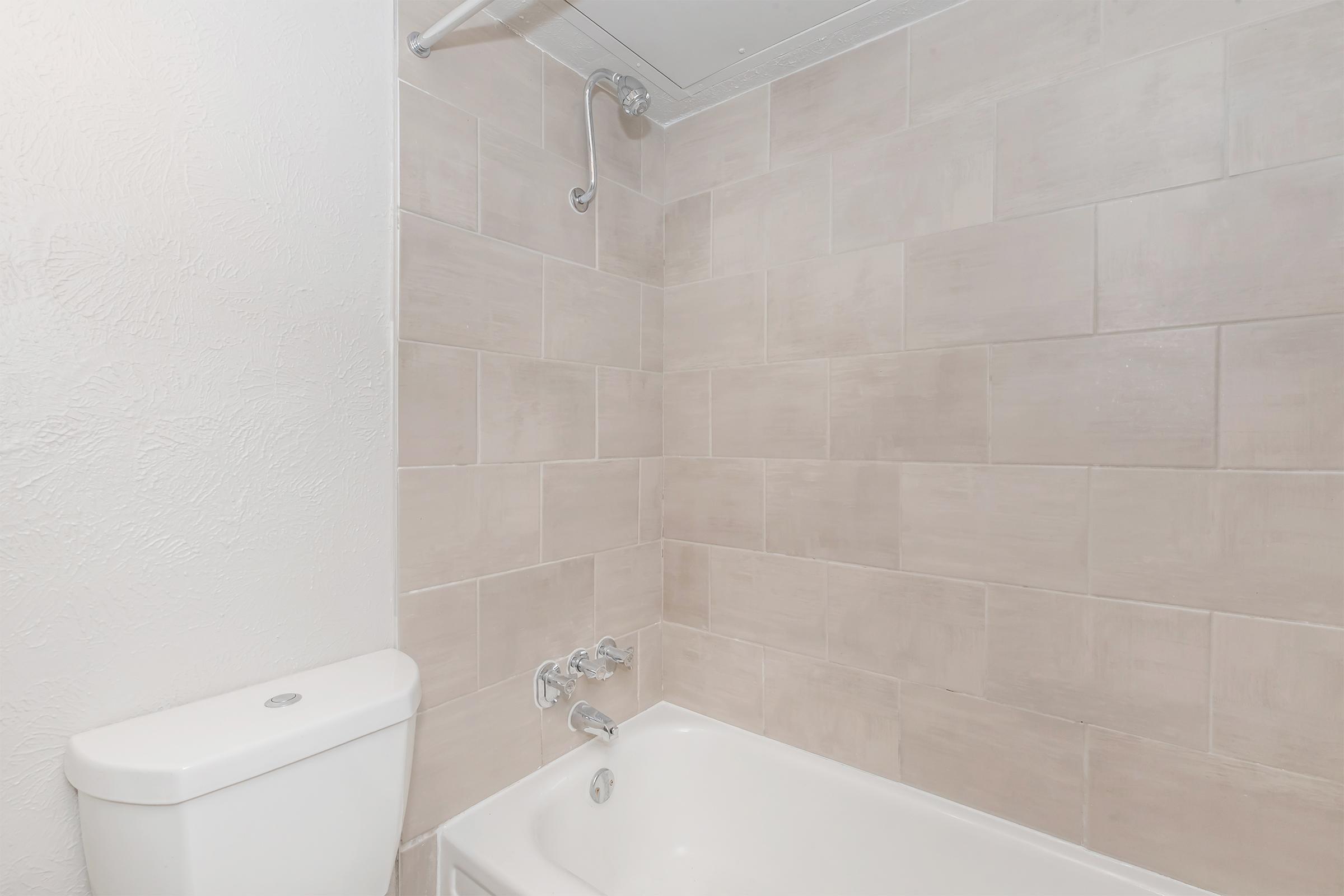 A clean bathroom featuring a modern bathtub with a showerhead above it. The walls are tiled in a light beige color, and there is a white toilet adjacent to the bathtub. The overall appearance is simple and contemporary.