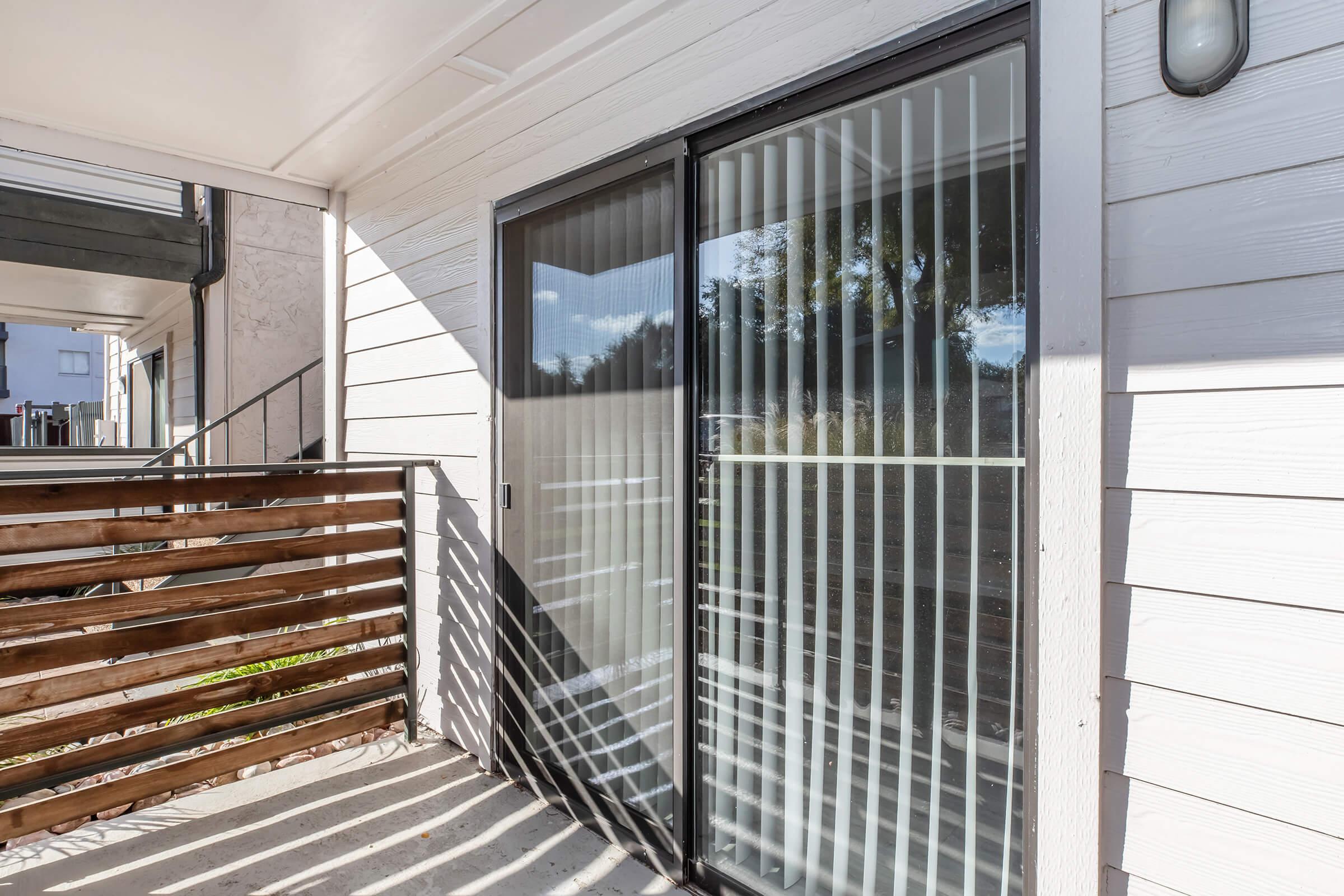 A well-lit balcony area featuring sliding glass doors with vertical blinds. Sunlight casts shadows on the floor, and a wooden railing is visible in the foreground, adding a touch of warmth to the modern exterior of the building.
