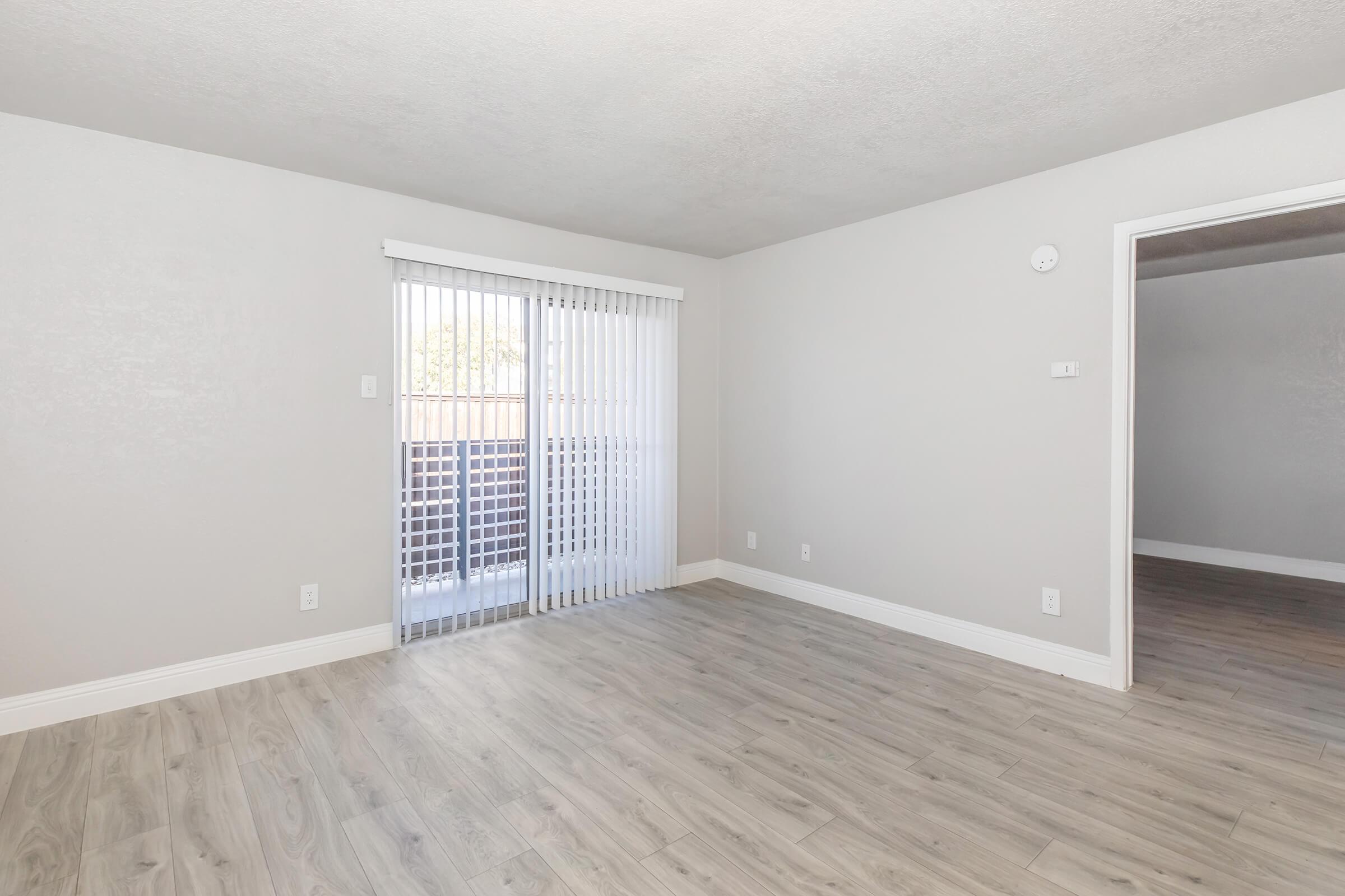 Empty room with light gray walls and wooden flooring. A large sliding glass door with vertical blinds allows natural light, leading to a balcony. The room features a simple design, with a wall switch and an additional door visible in the background. The overall ambiance is bright and airy.