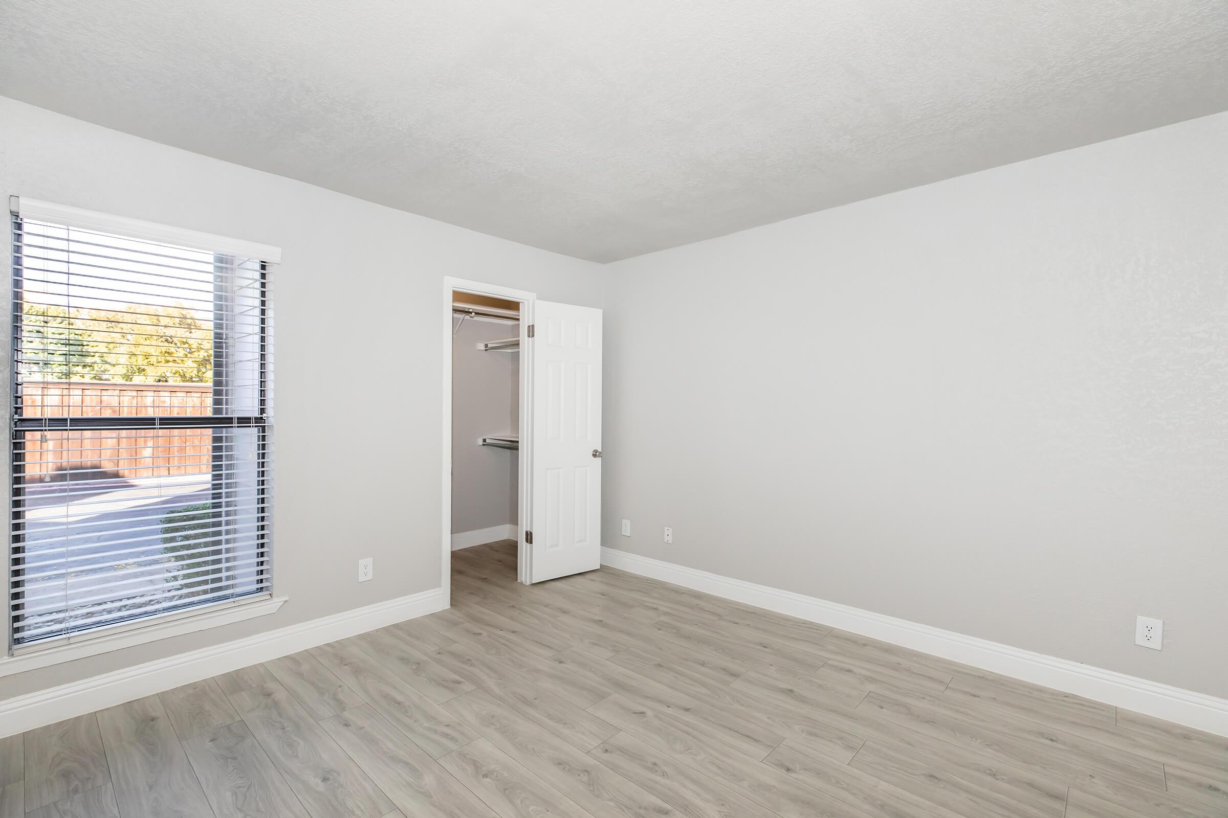 A well-lit room with light gray walls and a wood-like floor. There’s a window with horizontal blinds on one side, and a door leading to a small closet on the other. Natural light illuminates the space, creating a fresh and open atmosphere.