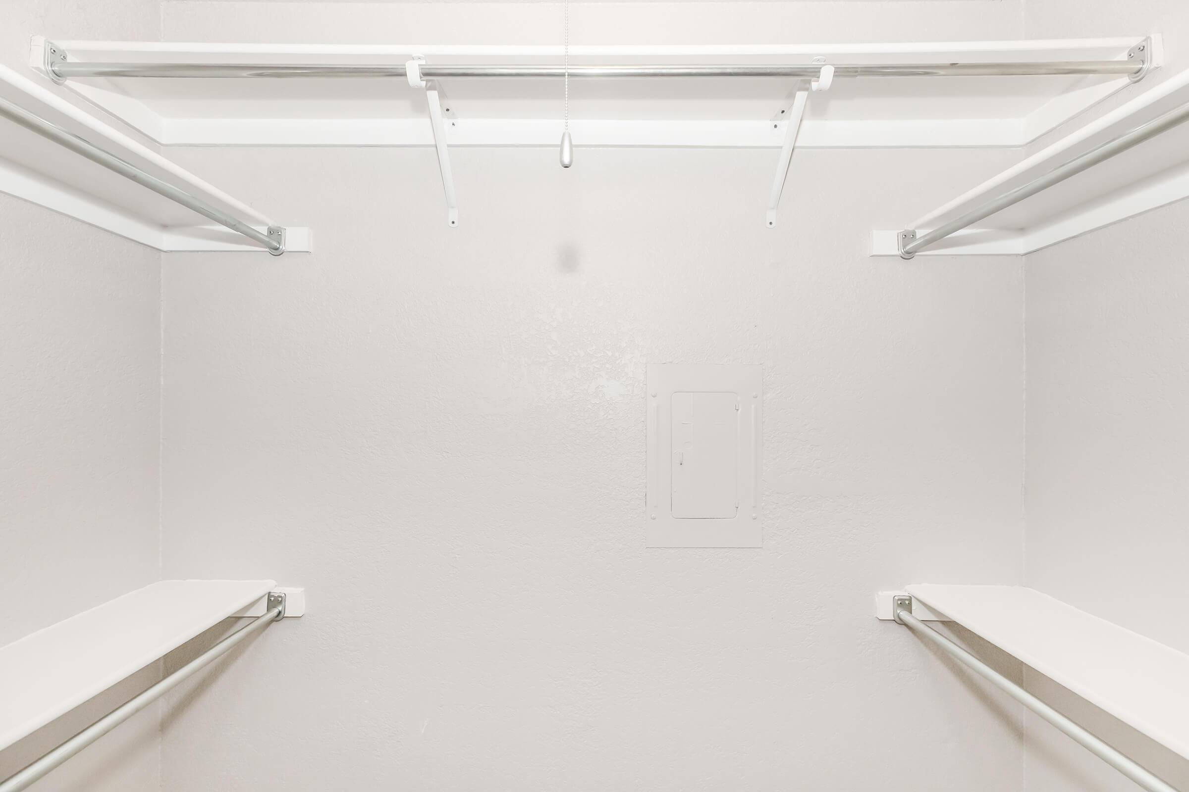Empty closet interior with light gray walls, featuring two parallel metal rods for hanging clothes and two shelves above, providing storage space. A small panel on the wall is present, indicating potential access for utilities or wiring.