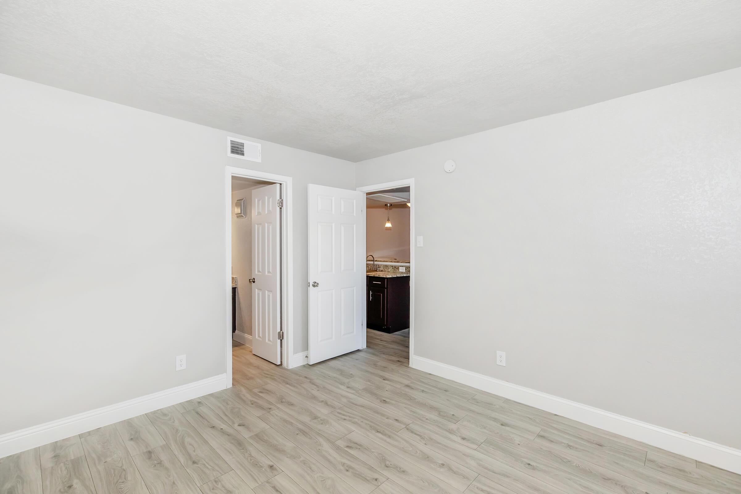 A spacious room with light gray walls and laminate flooring. It features two white doors: one leading to a bathroom and the other possibly to a kitchen area. The room is well-lit, enhancing the modern and clean ambiance. The simplicity of the design offers a neutral backdrop.
