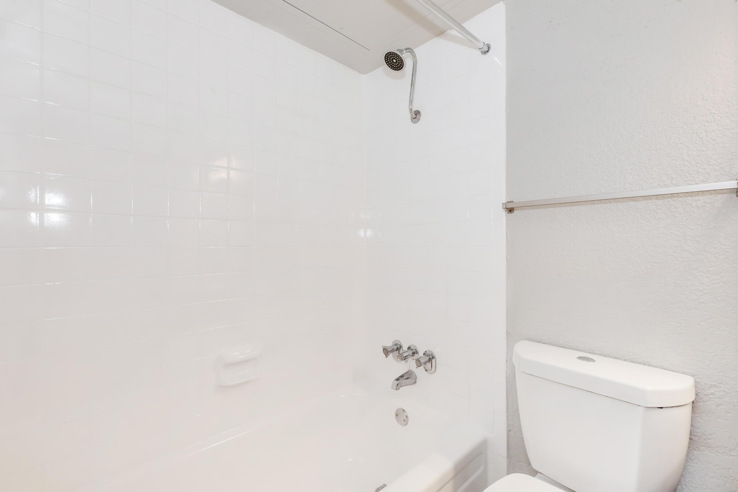 A clean, minimalistic bathroom featuring a white tiled shower with a showerhead, a bathtub, and a modern white toilet. The walls are light gray, and there is a towel rack mounted on the wall. The overall aesthetic is simple and bright.