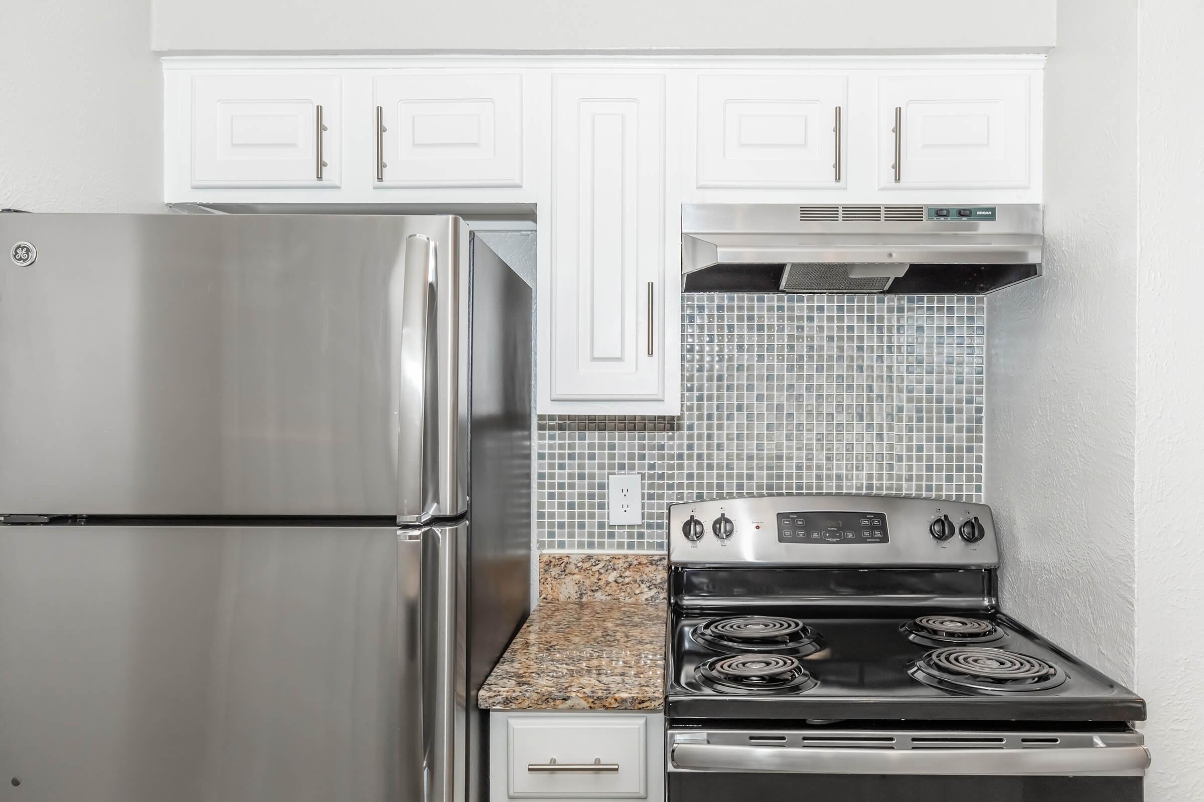 A modern kitchen featuring a stainless steel refrigerator, a black stove with four burners, and a silver range hood. The backsplash is made of small blue and gray tiles, and there are white cabinets above the countertop, which has a granite surface.