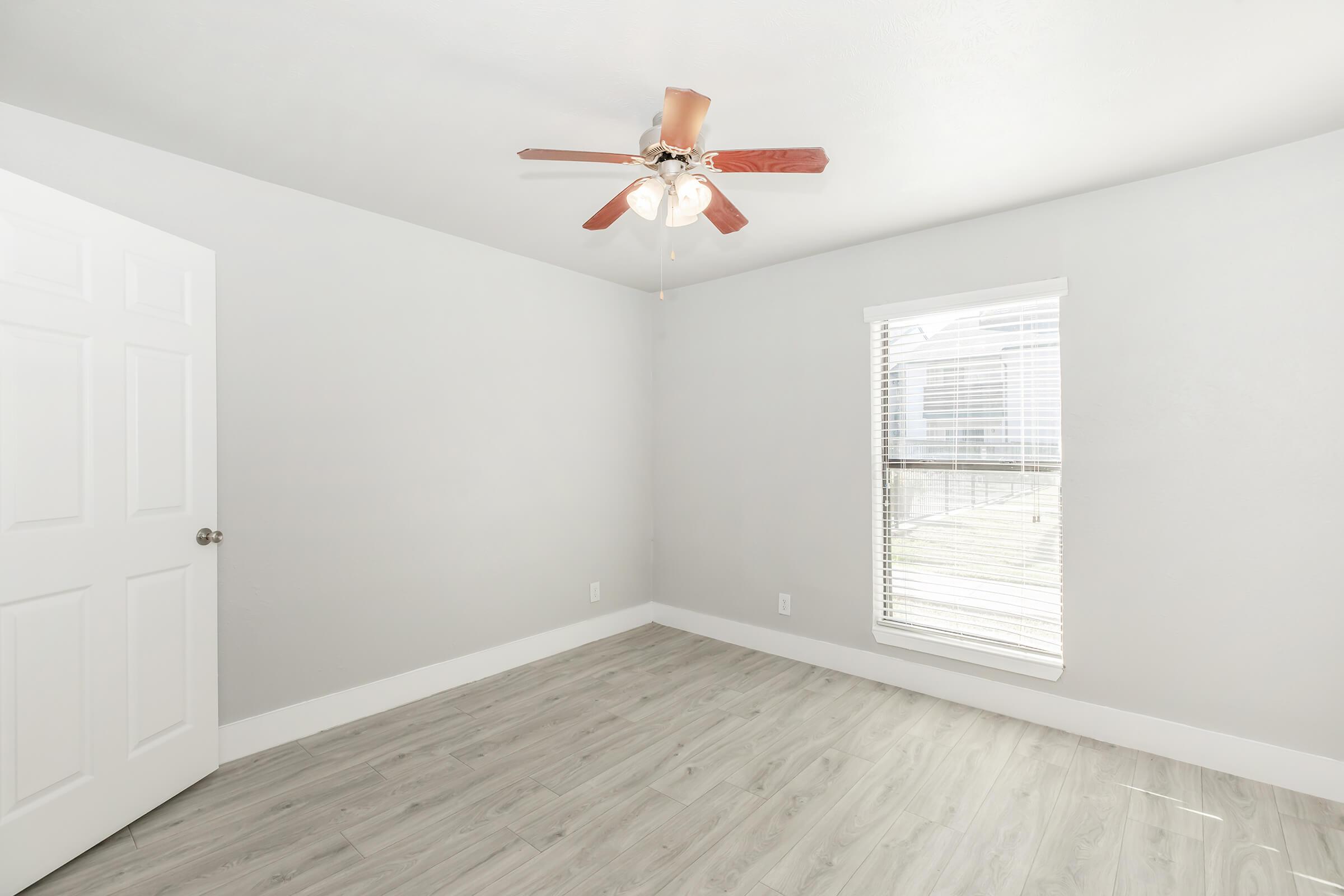 A light gray vacant room with a ceiling fan featuring wooden blades, a single white door on the left, and a window with blinds allowing natural light to enter. The floor is light-colored wood, and the walls are neutral-toned, creating a clean and spacious atmosphere.