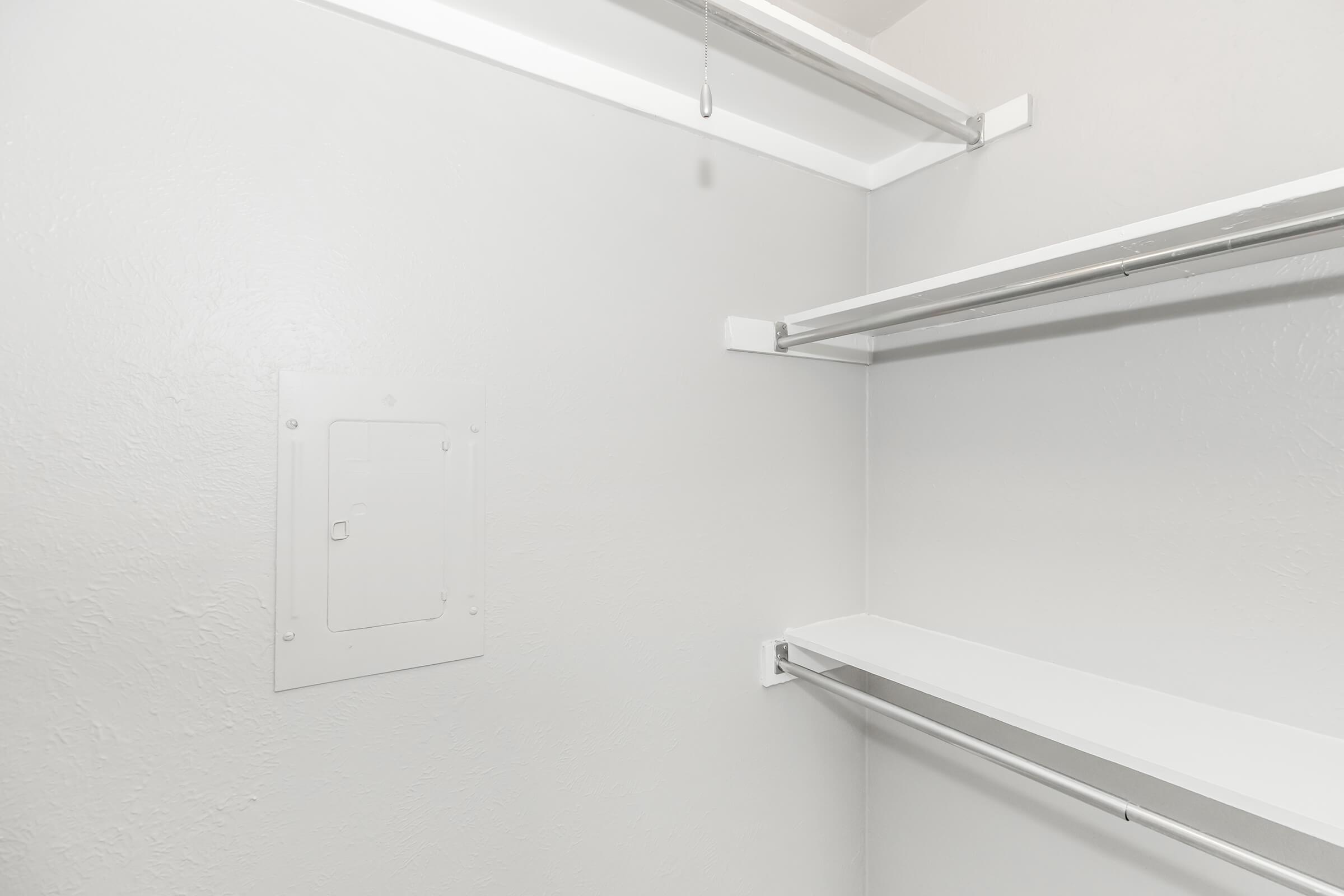 A clean, empty closet with white walls. Two horizontal shelves are mounted on opposite sides, providing storage space. A small access panel is visible on the left wall. The overall atmosphere is bright and minimalistic.
