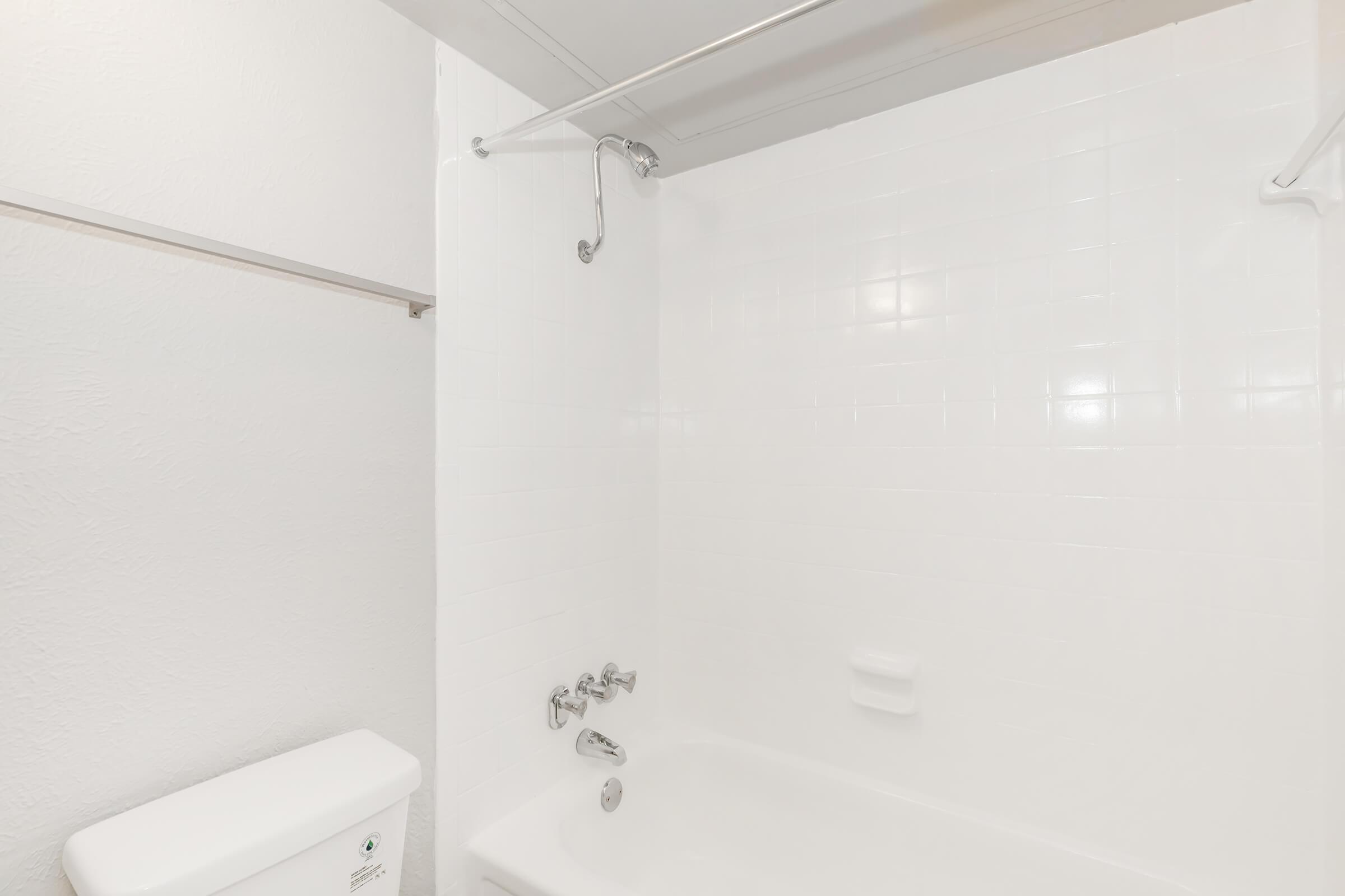 A clean, simple bathroom featuring a white bathtub, wall-mounted showerhead, and chrome fixtures. The walls are tiled in a glossy white finish, and a toilet is positioned beside the bathtub. The overall aesthetic is bright and minimalist, reflecting a modern design.