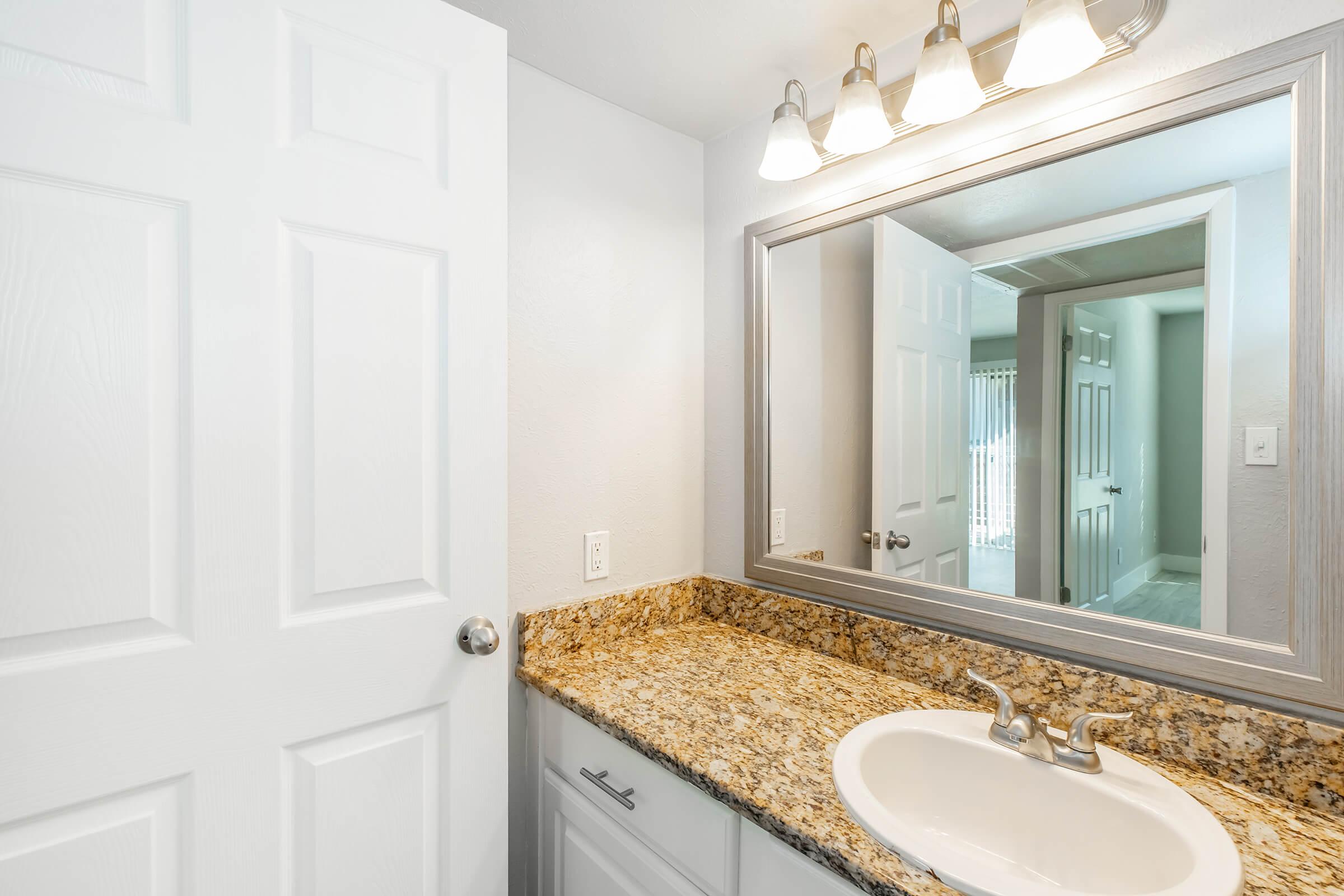 A well-lit bathroom featuring a white door, a large mirror above a granite countertop with a sink, and ceiling lights. In the background, there are doors leading to other rooms and a window allowing natural light. The overall color scheme is light and neutral, creating a clean and modern appearance.