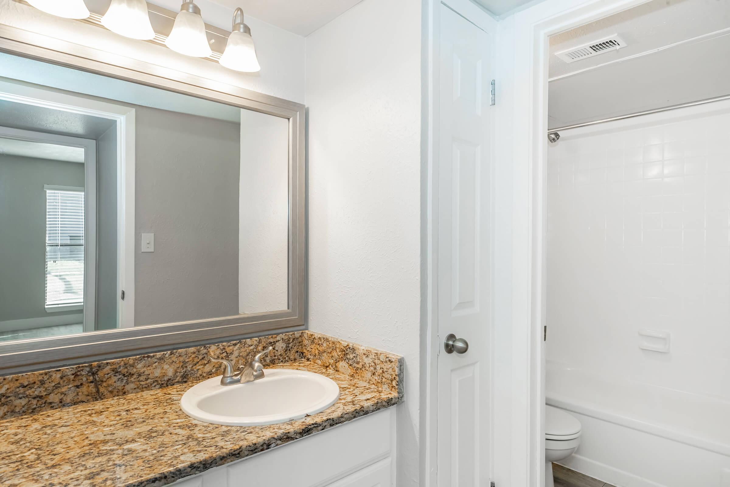 A bright bathroom featuring a granite countertop with a sink and a large mirror above it. The wall beside the mirror is painted white, and there is a shower area with a sliding door visible in the background. Natural light filters in through a nearby window, enhancing the airy feel of the space.