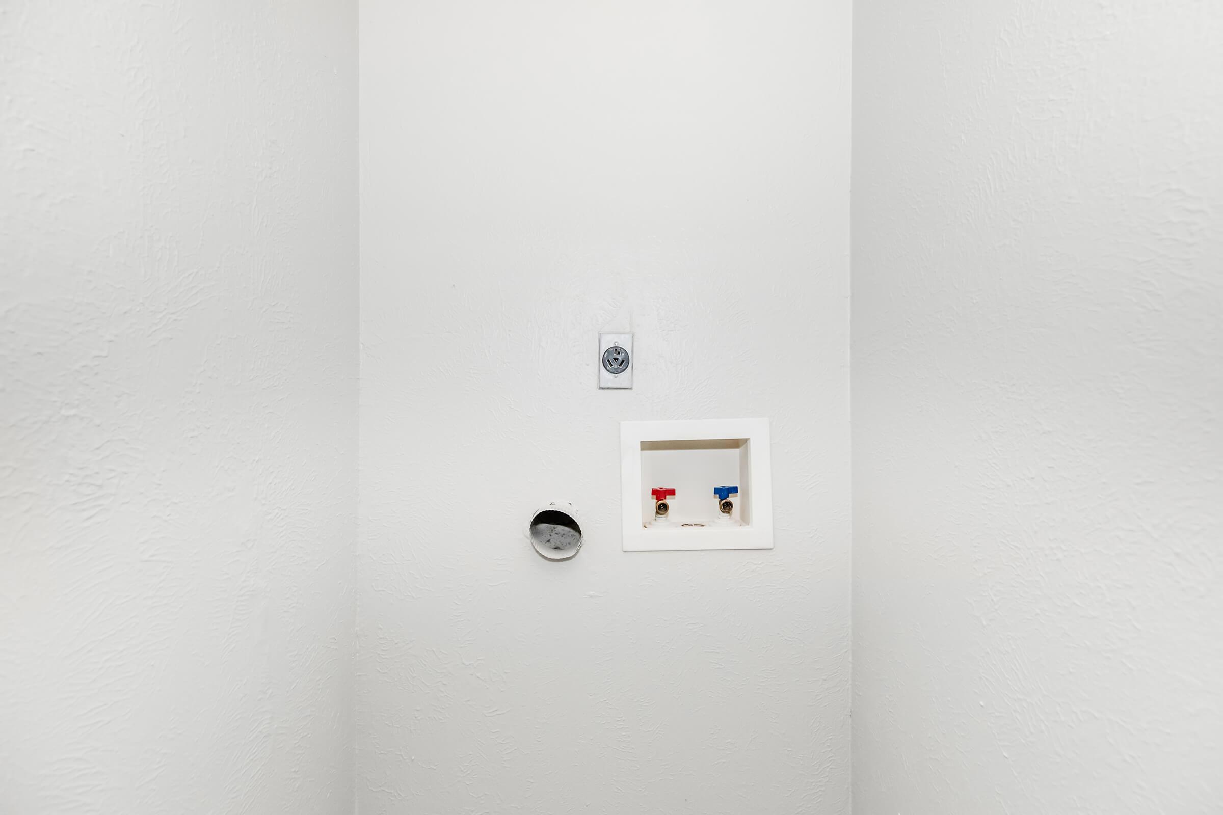 A minimalist, white-walled space featuring a small wall-mounted electrical outlet, a round hole in the wall, and a recessed fixture with red and blue knobs, likely for plumbing or utility access. The overall aesthetic is clean and simple, emphasizing the neutral color scheme.