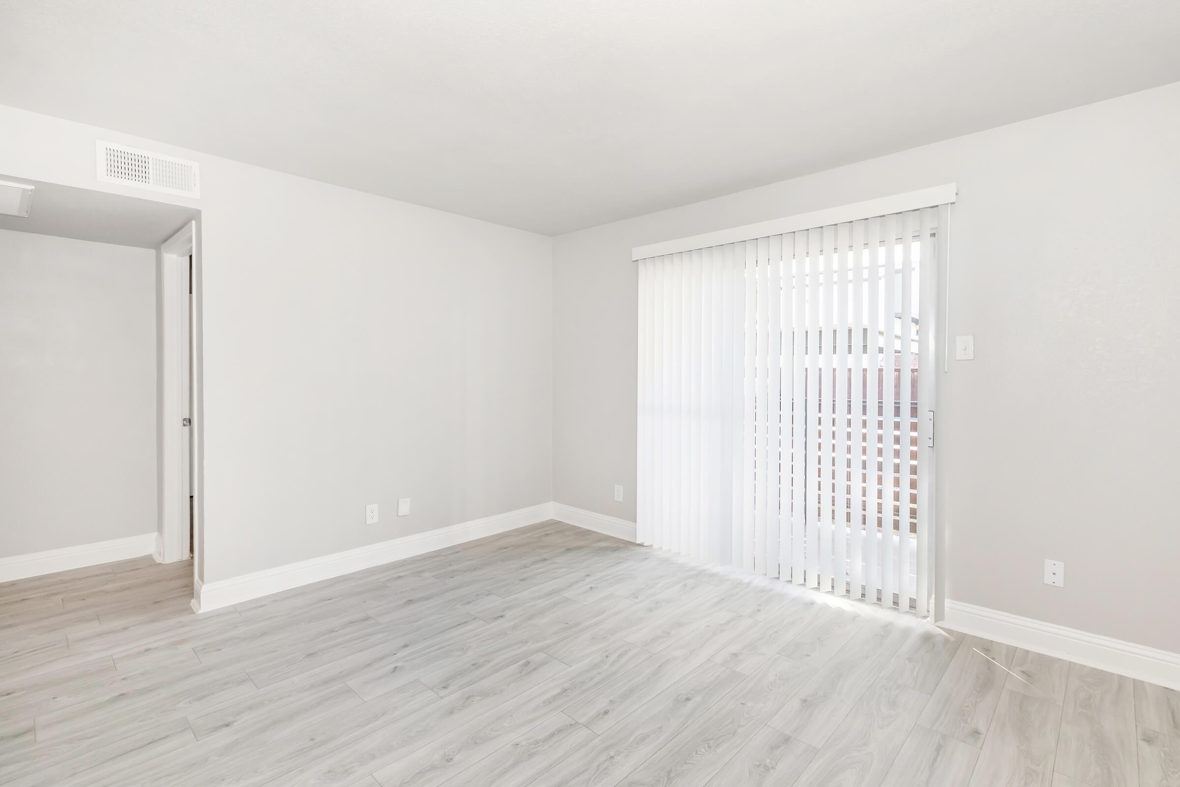 Empty room with light gray walls and light wood-like flooring. A sliding glass door with vertical blinds allows natural light to enter. There is a doorway to the left, leading to another space. Overall, the room has a modern and minimalistic aesthetic.