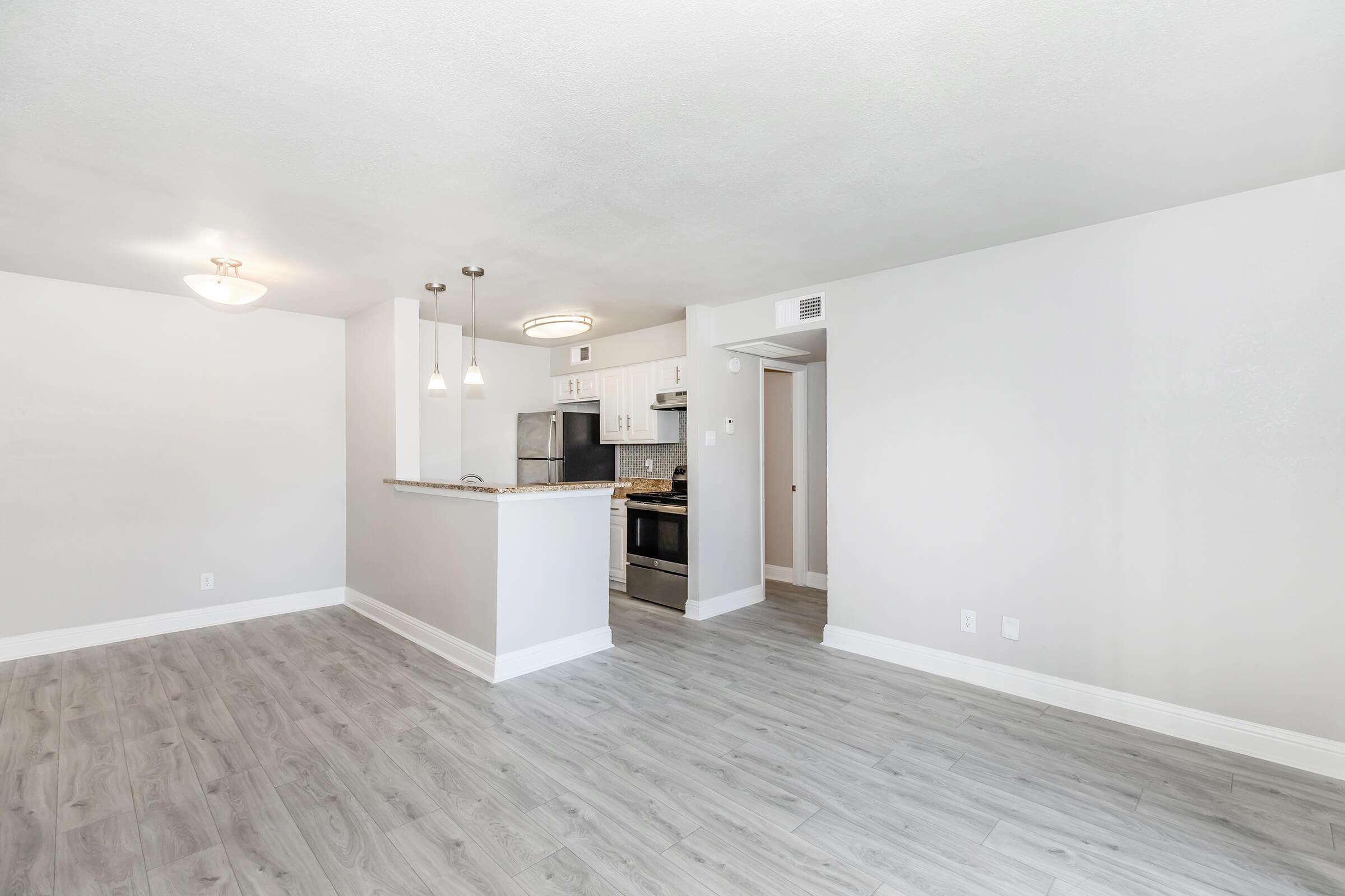 A spacious, modern living area with light gray walls and wood-like flooring. The kitchen features a breakfast bar with pendant lighting, stainless steel appliances, and ample cabinetry. Natural light fills the room, highlighting the open layout and inviting atmosphere.