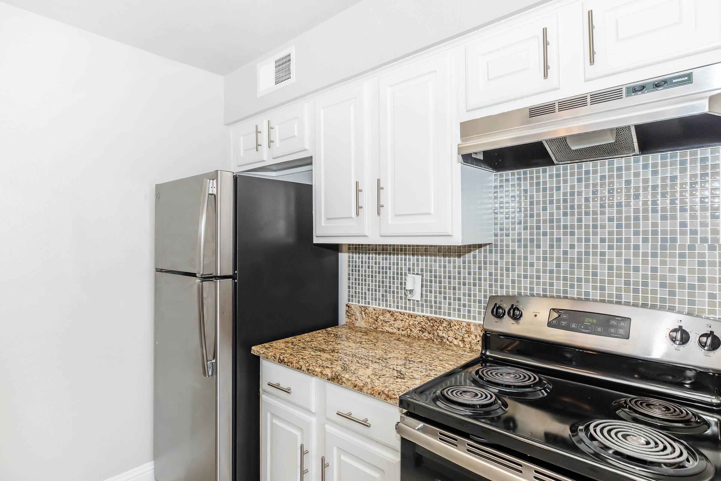 A modern kitchen featuring a black refrigerator, a stainless steel oven with a smooth top, white cabinets, and granite countertops. The backsplash consists of small blue tiles, creating a clean and stylish look.