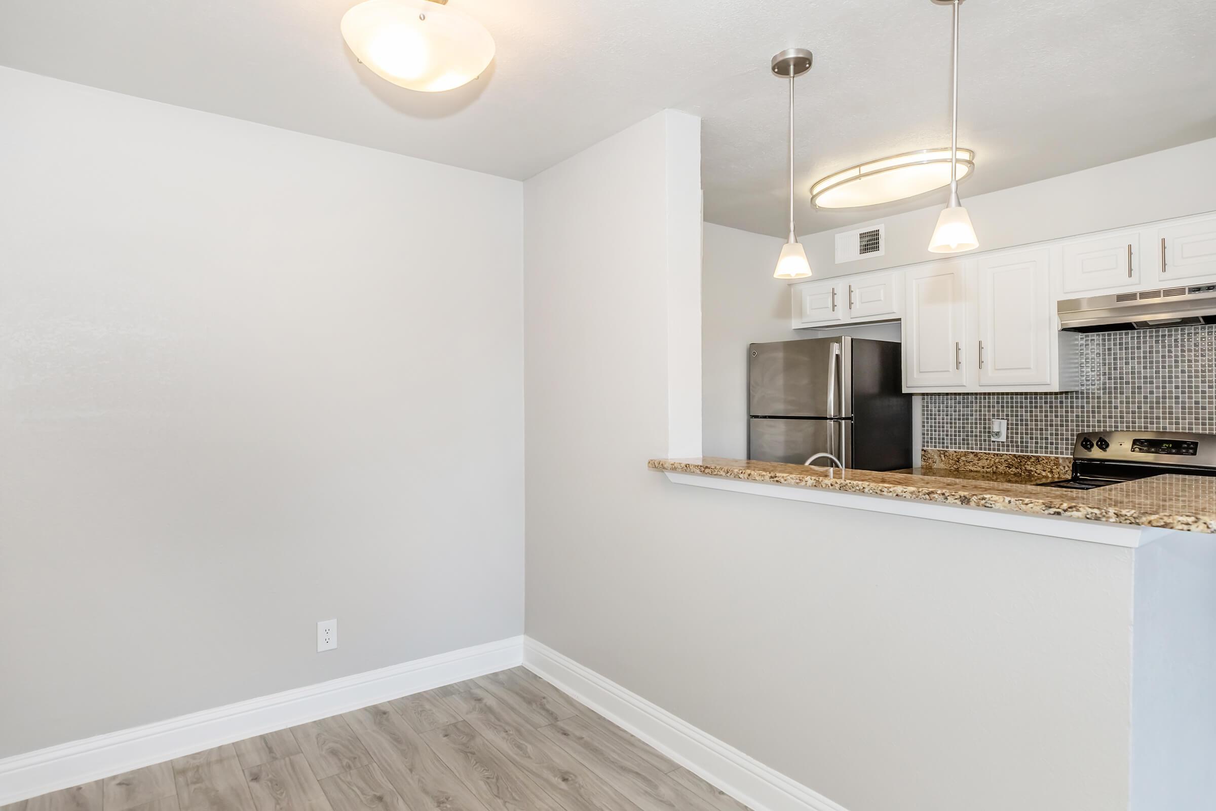 A modern kitchen area featuring a granite countertop with bar seating, stainless steel appliances, and white cabinets. The walls are painted in a light color, and there are two pendant lights hanging from the ceiling, providing ample lighting in the space.