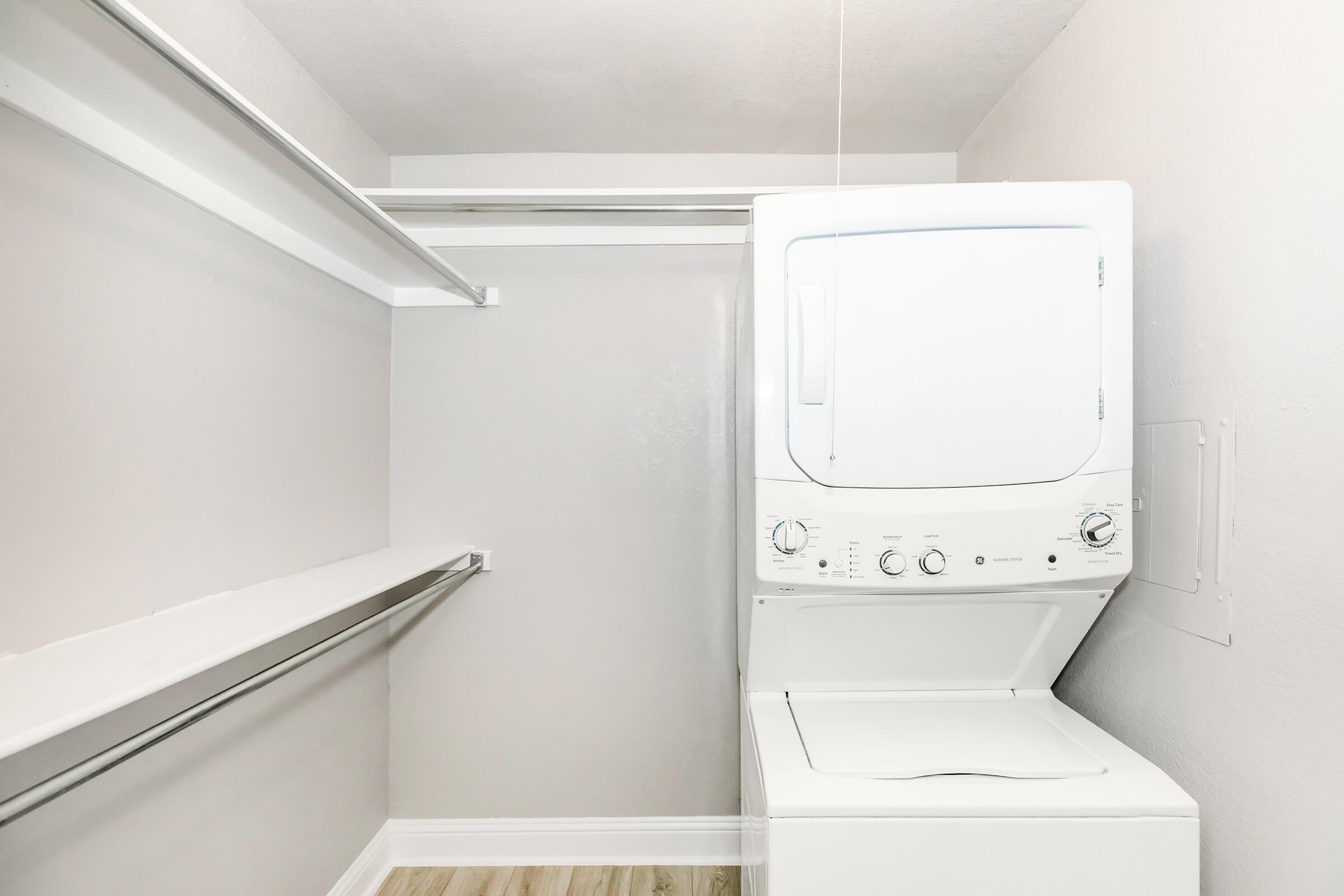 A compact laundry area featuring a stacked washer and dryer against a light gray wall, with two ceiling-mounted shelves for additional storage. The floor is a light wood finish, and the space is organized and tidy, making it ideal for efficient laundry management.