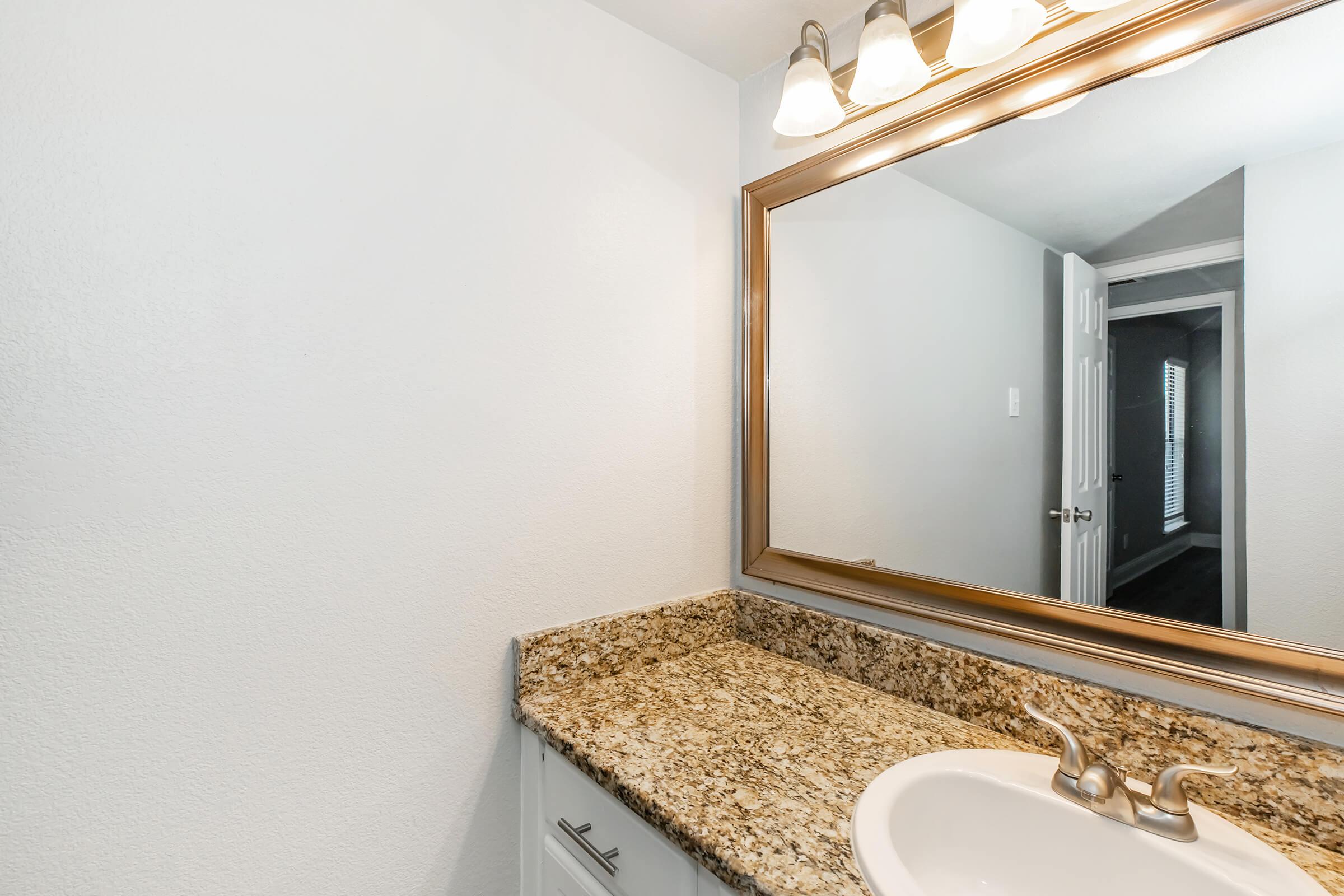 A well-lit bathroom featuring a granite countertop with a white sink, a large mirror above the sink, and modern light fixtures. In the background, a door is visible, leading to another room. The walls are painted white, creating a clean and simple aesthetic.