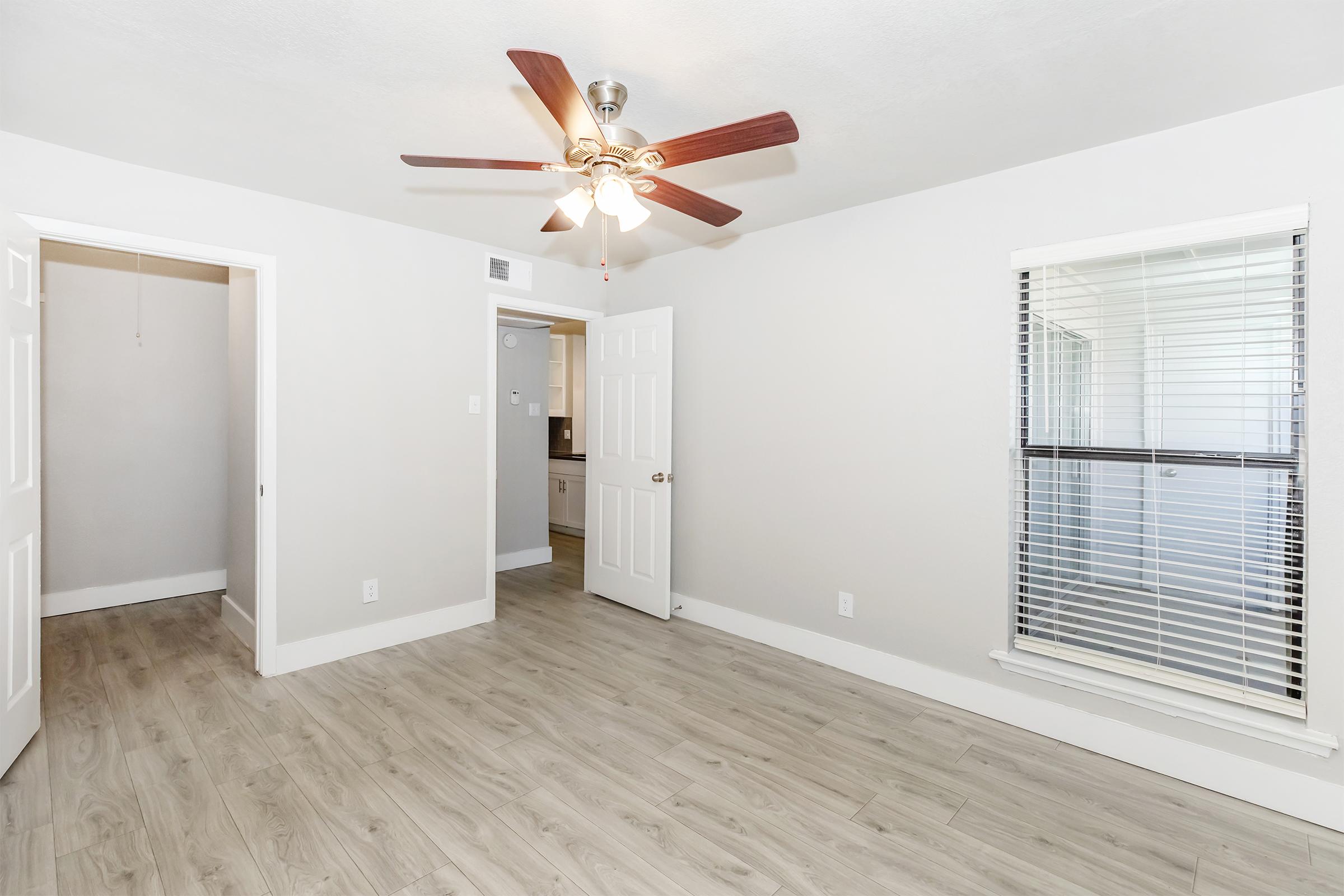 A bright room featuring a ceiling fan with wooden blades, light-colored walls, and laminate flooring. It includes a window with blinds, a doorway leading to another room, and an open space that connects to a kitchen area. The overall ambiance is clean and inviting.