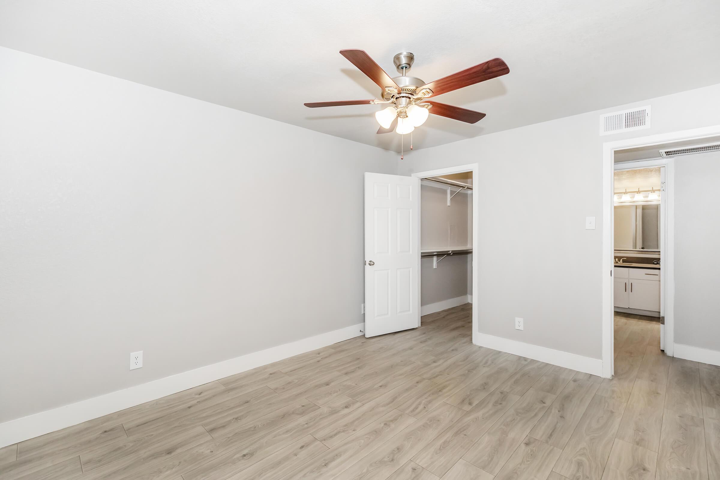 A spacious room with light gray walls and wood-style flooring. A ceiling fan with light hangs in the center. To the right, there's an open door leading to a closet, and in the background, another door opens to a bathroom area. The ambiance is bright and inviting.