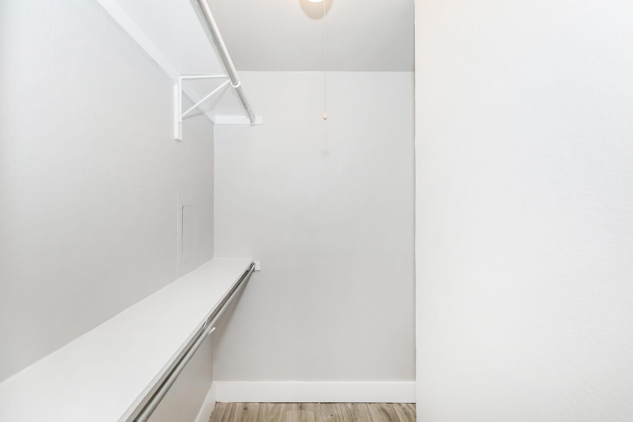 A spacious, empty closet featuring white walls, a wooden floor, and a simple overhead light fixture. There are shelves along one side and a long rod for hanging clothes, highlighting the room's minimalistic design. The overall appearance is bright and uncluttered.