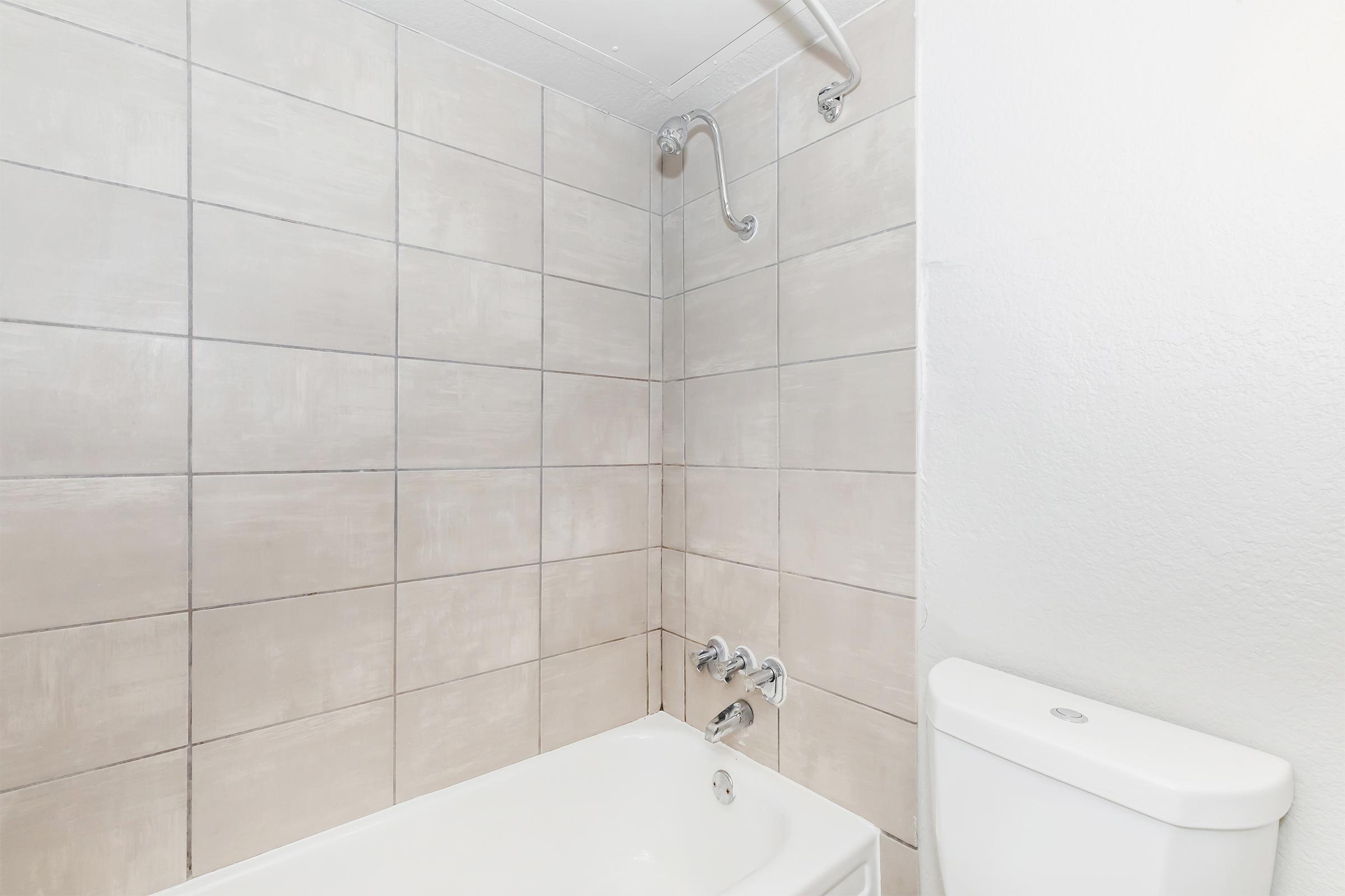 A modern bathroom featuring a white bathtub with a chrome faucet and showerhead. The walls are tiled in light gray with a contemporary design. A white toilet is visible to the right, complementing the clean and minimalistic aesthetic of the space.