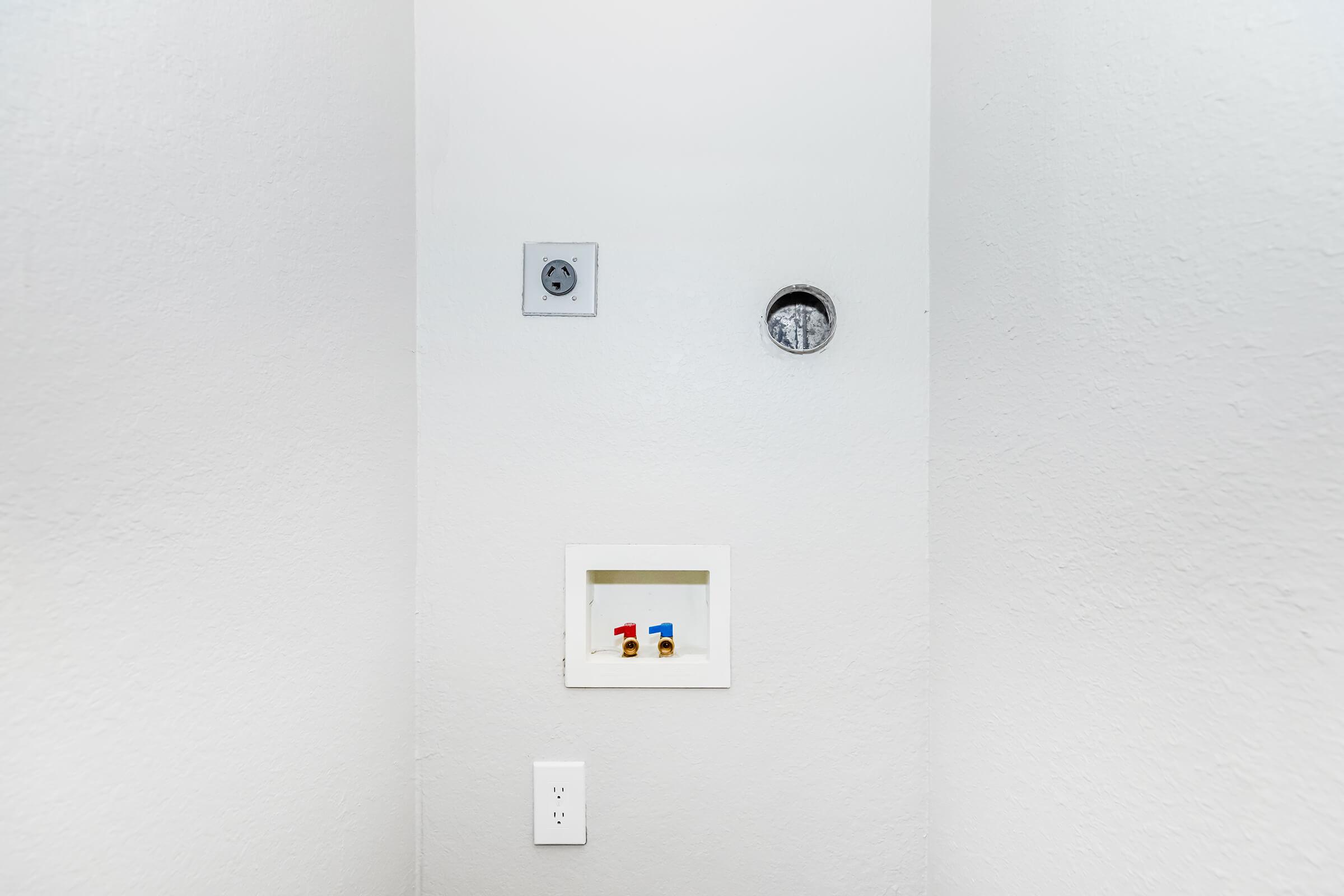 A simple gray wall with a small rectangular opening at the bottom showing colorful items inside. Above, there is a circular hole and an electrical outlet on the left side. The overall space appears clean and minimal, emphasizing the neutral tones of the wall.