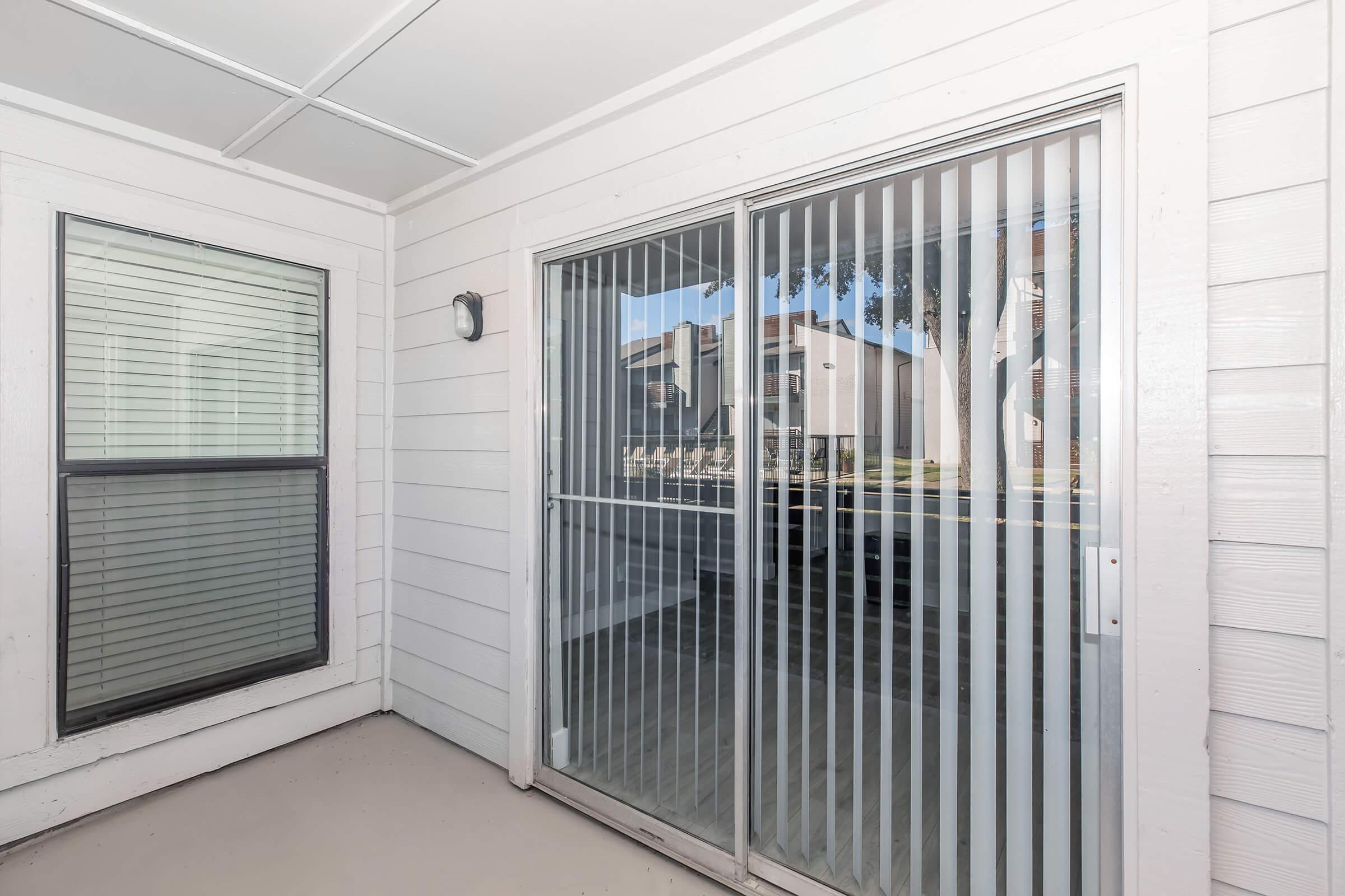 A covered patio area featuring a sliding glass door with vertical blinds, alongside a window with horizontal blinds. The wall is white, and the view outside shows neighboring buildings and greenery. The space appears clean and well-lit, suggesting a comfortable outdoor area.