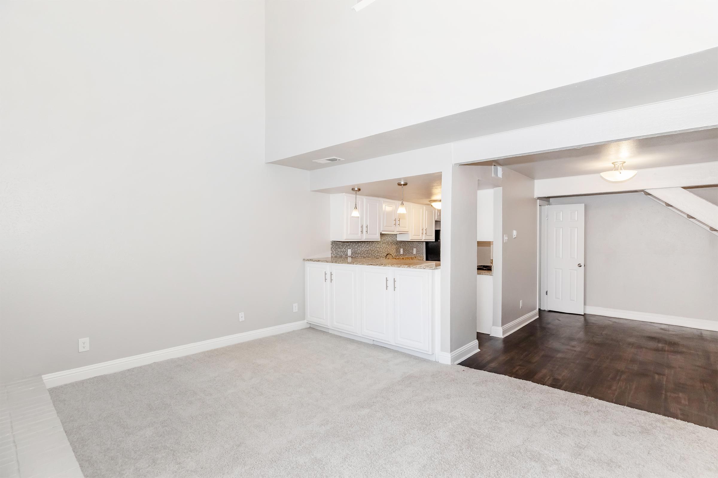 A spacious, modern living area with light gray walls and plush carpeting. To the left, a kitchen with white cabinetry and granite countertops is partially visible. A doorway leads to another room, and the room features high ceilings, creating an airy feel. Natural light enhances the open space.