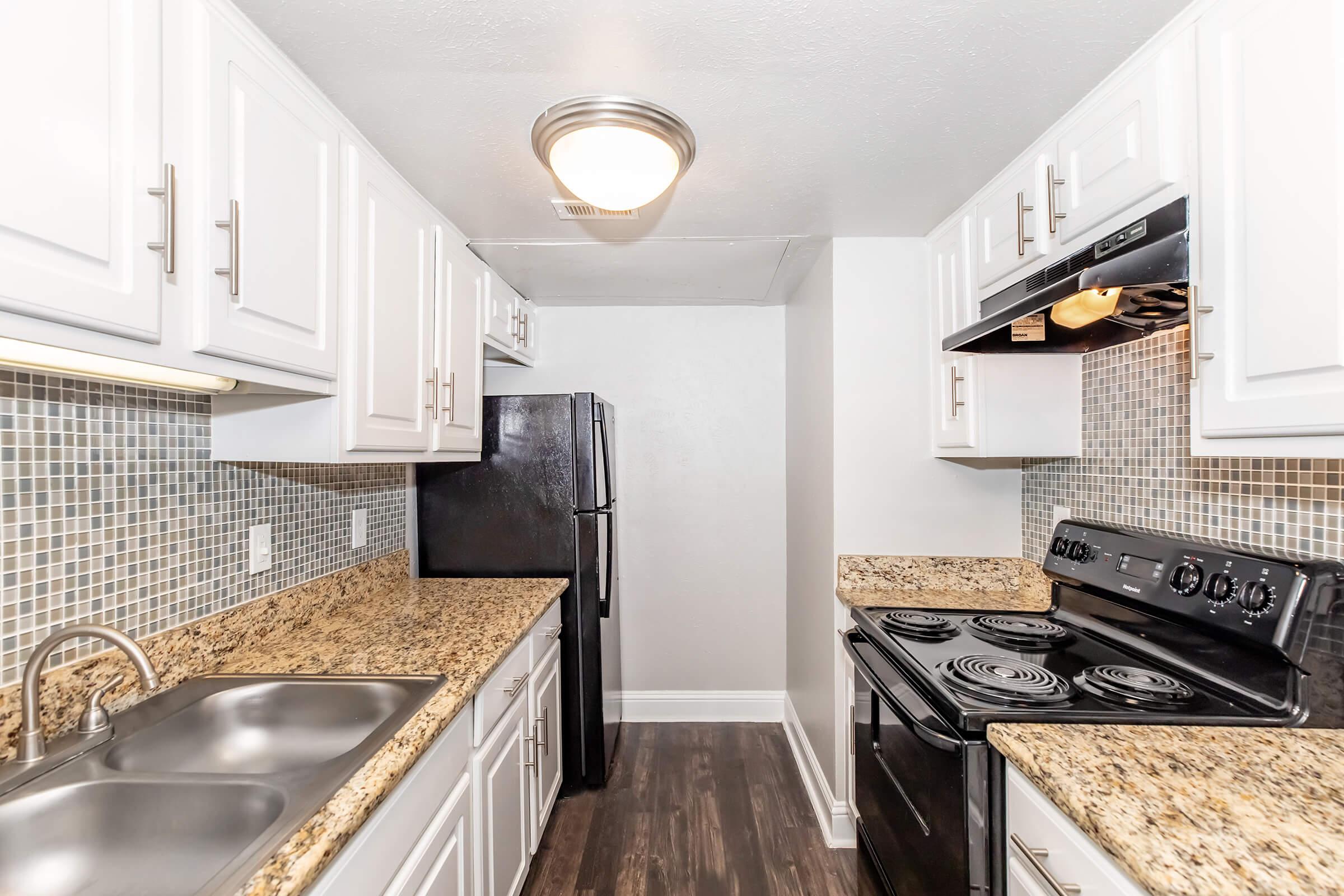 A modern kitchen featuring white cabinets, granite countertops, a double sink, stainless steel appliances, and black kitchen appliances. The backsplash is composed of small mosaic tiles, and there is a ceiling light fixture providing illumination. The flooring is dark and adds contrast to the overall design.