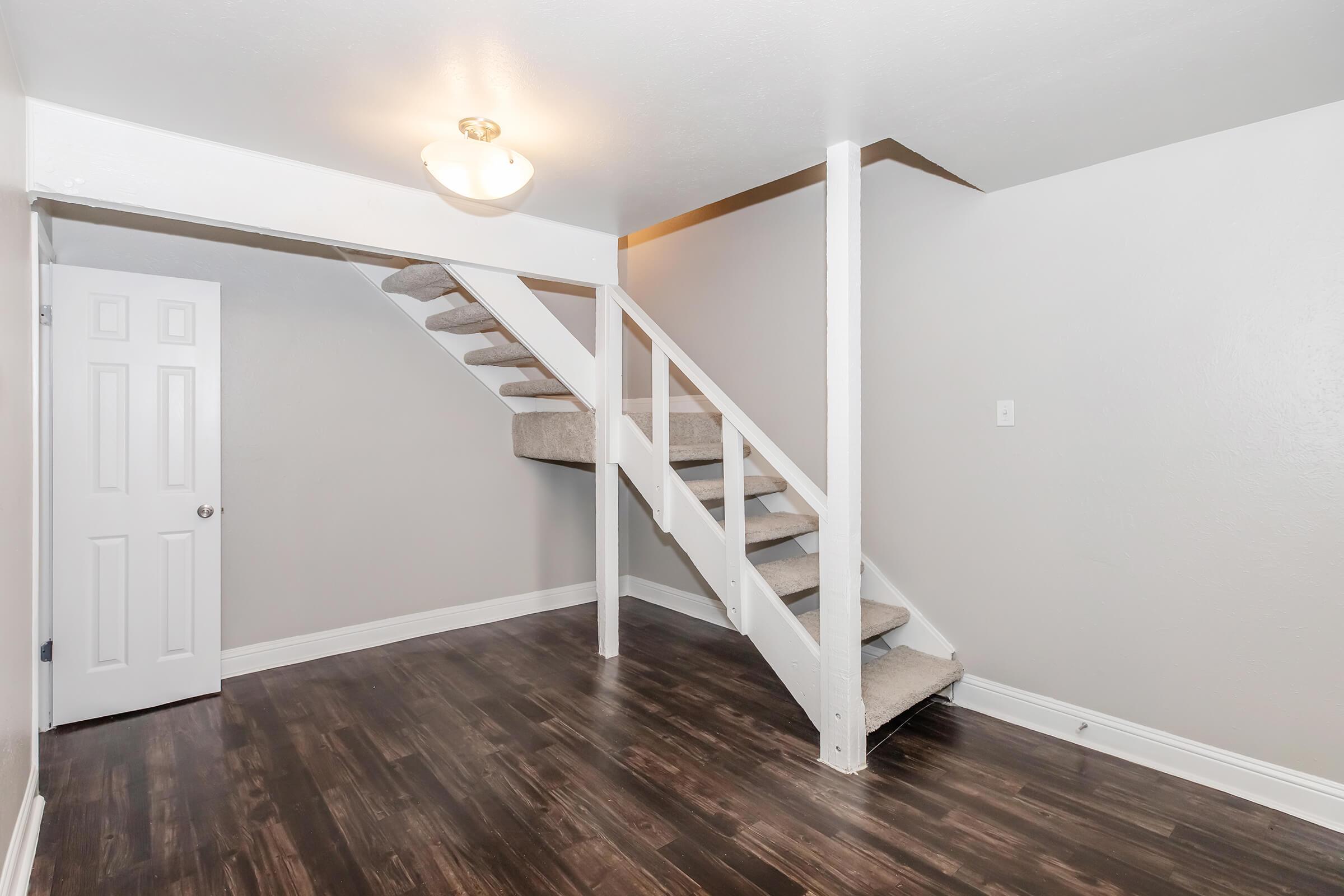 Interior view of a room featuring a staircase leading to a second level. The walls are painted a light grey, and the floor is dark wood. A door is visible on the left, and a ceiling light fixture hangs above. The space appears well-lit and clean, with carpeted steps on the staircase.