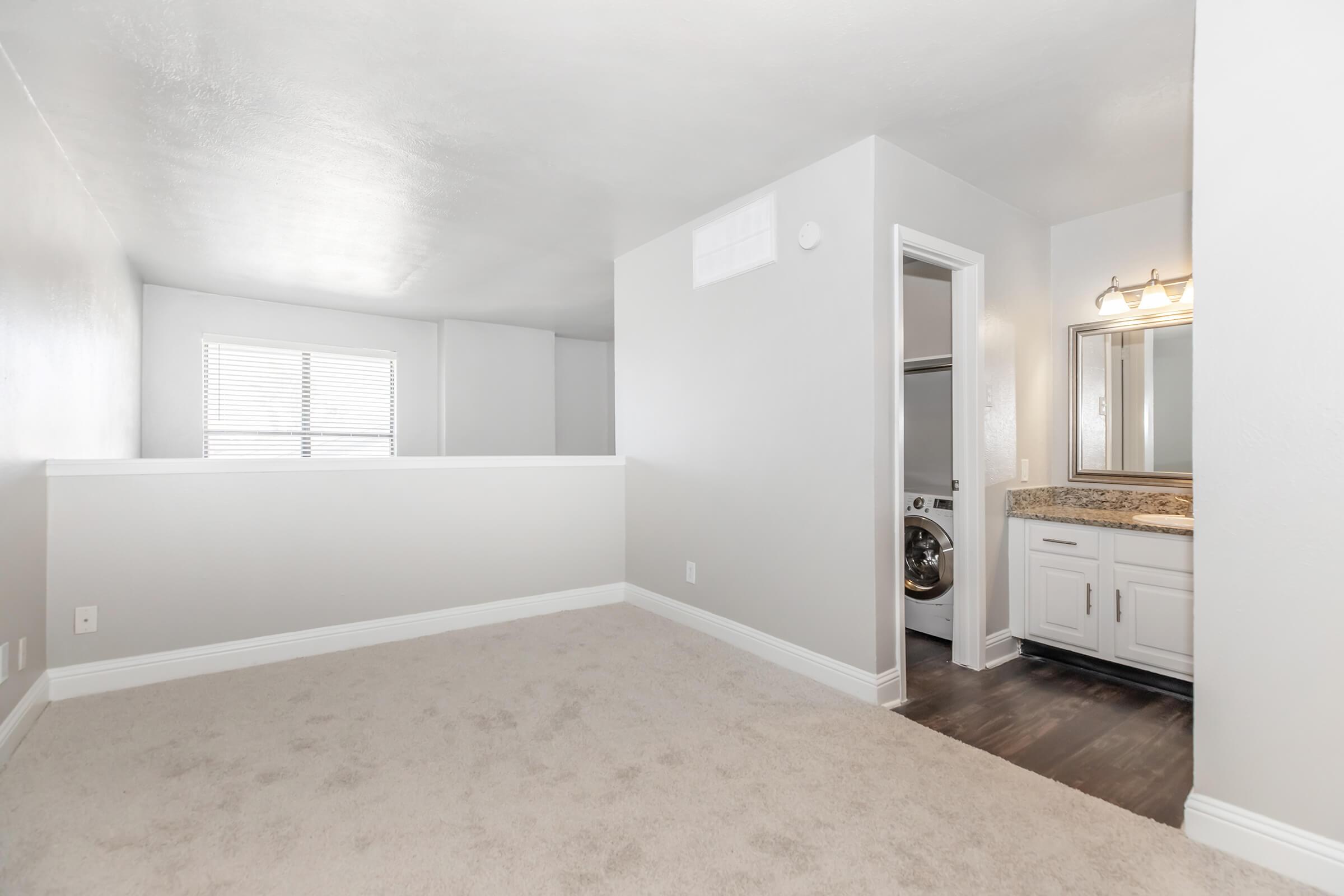 A spacious, well-lit room with light gray walls and plush carpet. On one side, there’s a window allowing natural light in, and on the opposite side, a doorway leads to a bathroom featuring a large mirror and a laundry area. The overall aesthetic is modern and clean, with a neutral color palette.