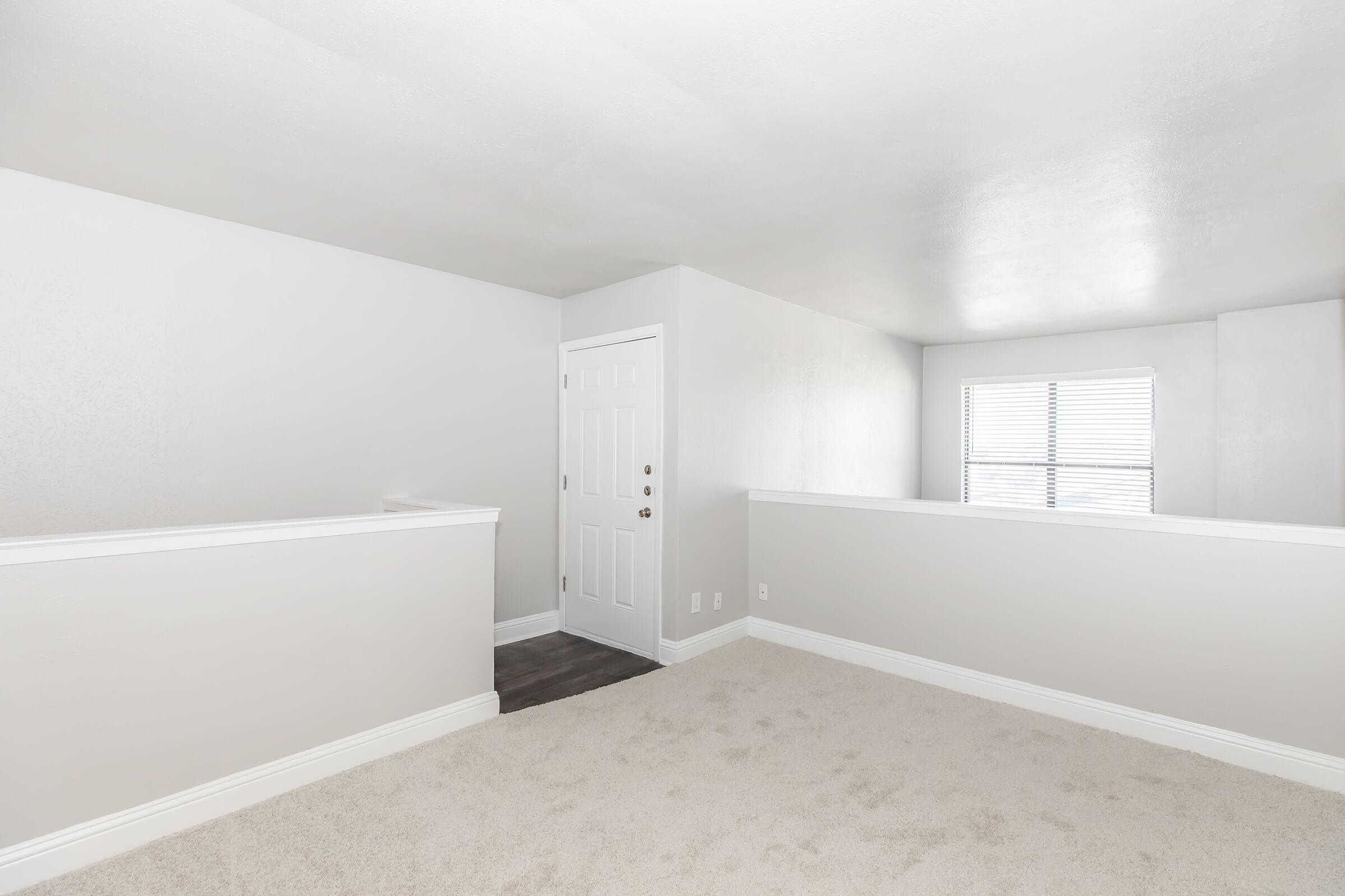 A spacious, empty room with light gray walls and soft beige carpet. There is a white railing along the left side and a doorway with a white door on the right. A window provides natural light, illuminating the room's minimalist design, creating a bright and airy atmosphere.