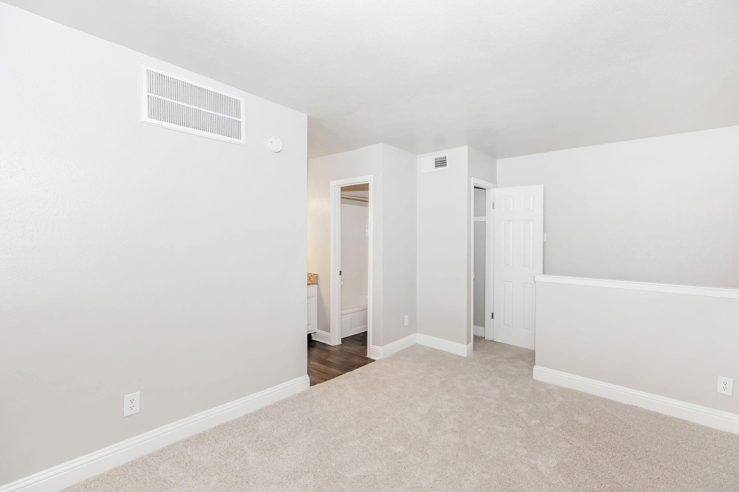 A well-lit, empty room with light gray walls and plush carpet. The space features a white door leading to another room and a vent in the ceiling. There is a small wall section with a railing, and the overall atmosphere appears clean and modern, suitable for various purposes.