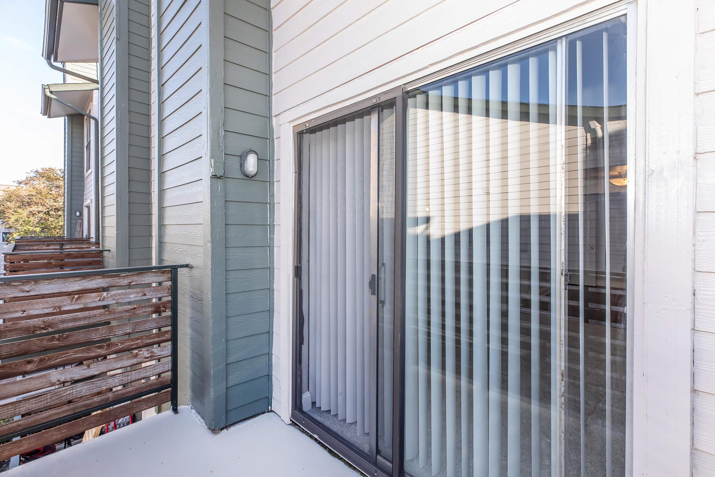 View of a balcony area featuring a sliding glass door with vertical blinds, leading to a spacious outdoor space. The exterior walls are partially painted in light colors, and a wooden railing adds an inviting touch. The scene is well-lit and showcases a modern design.