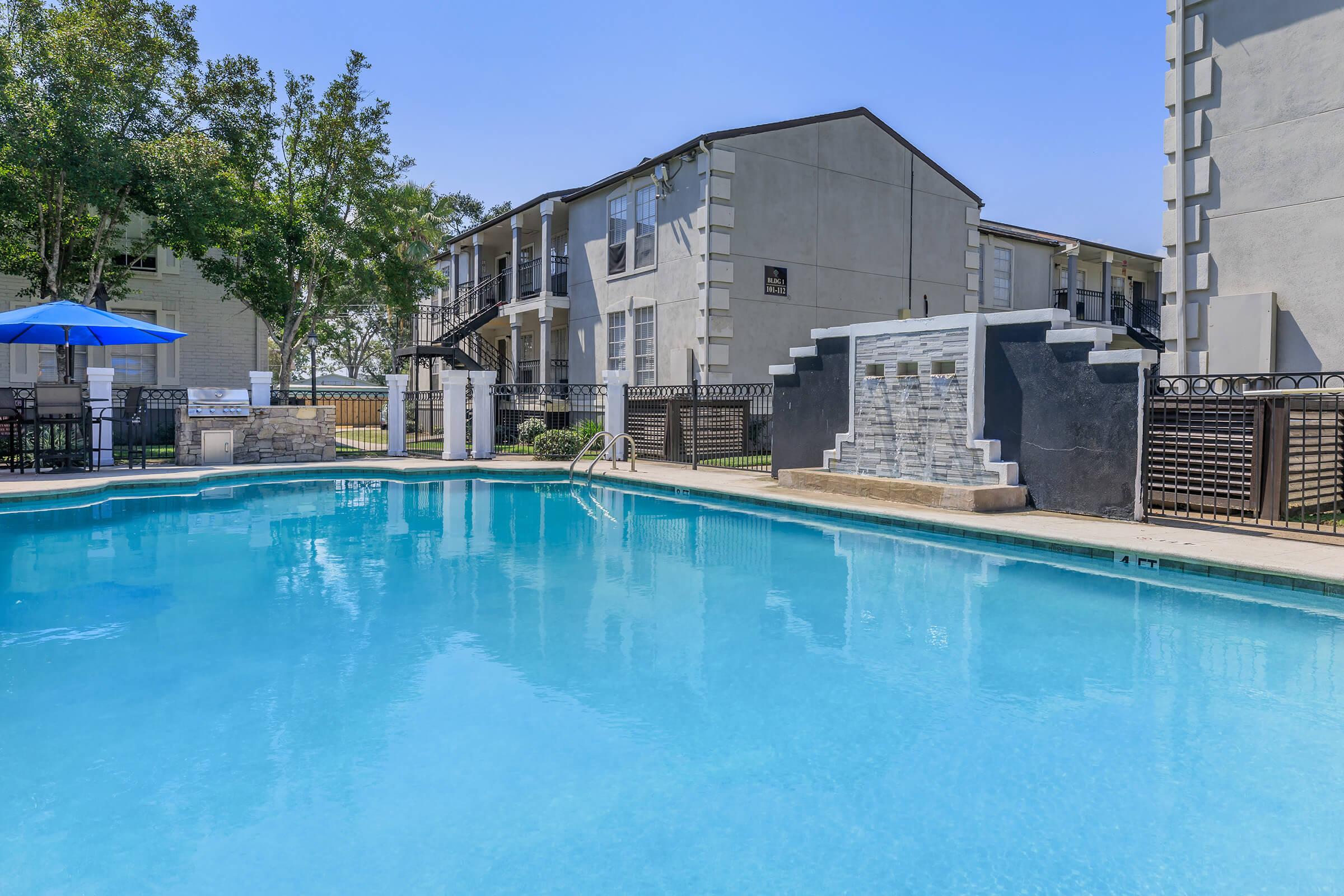 A clear blue swimming pool surrounded by a patio, featuring lounge chairs and an umbrella. In the background, an apartment building with multiple floors is visible, alongside landscaped trees and grass. The scene is sunny and inviting.