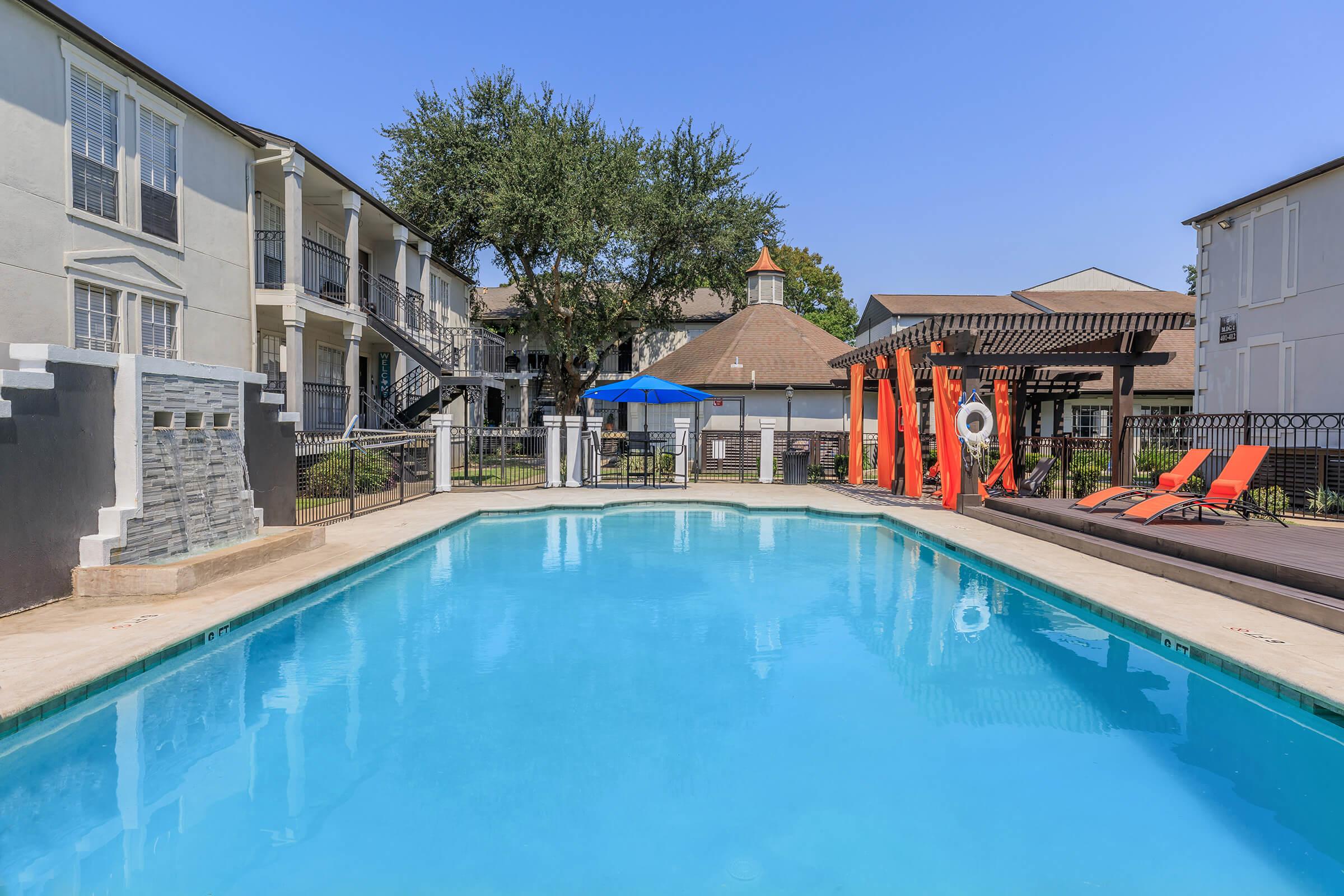 A bright and inviting swimming pool surrounded by lounge chairs and a shaded area with a pergola. The pool area is flanked by residential buildings with balconies, and a few trees provide a touch of greenery. Clear blue skies enhance the relaxing atmosphere of the outdoor space.