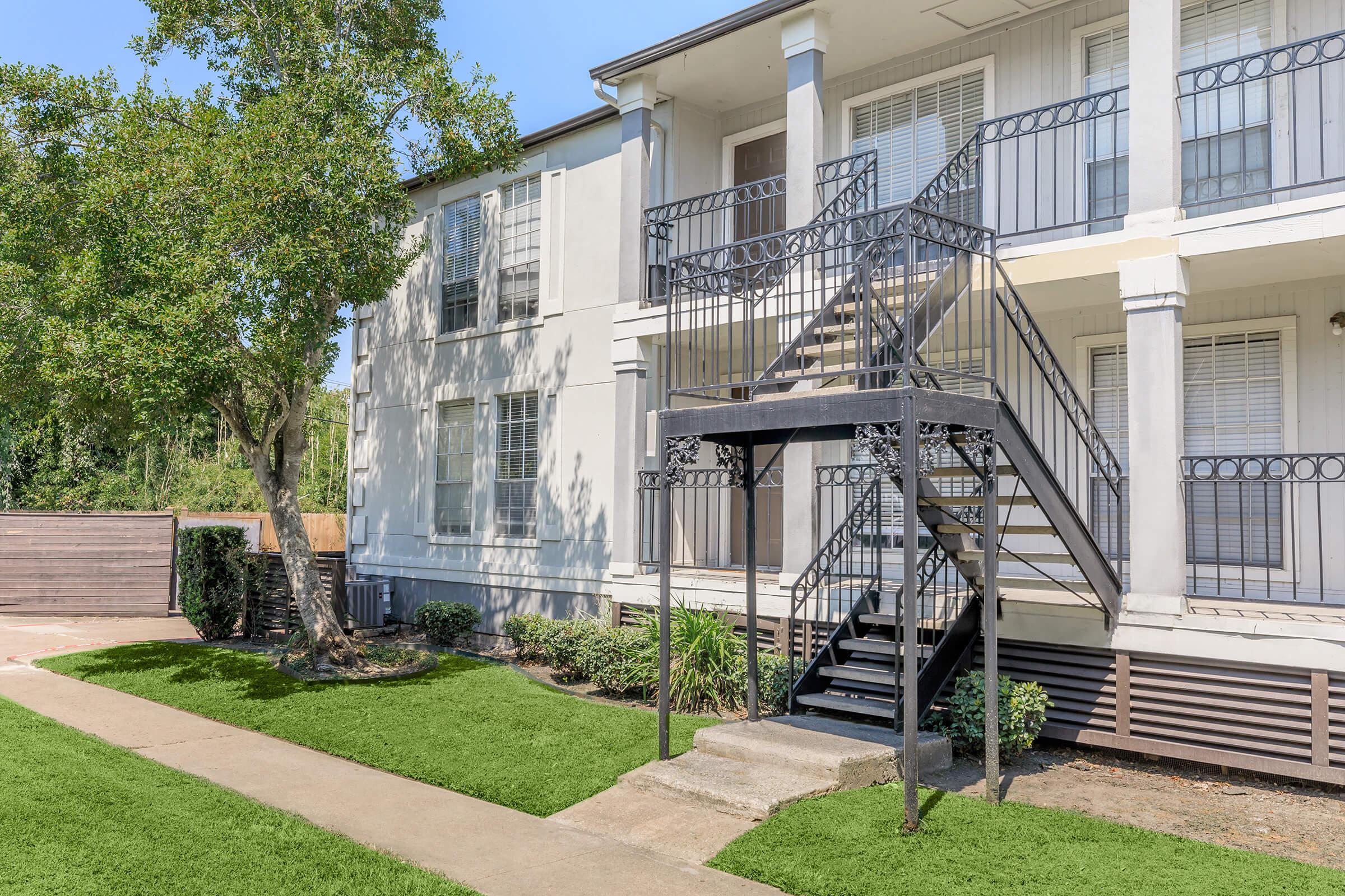 A two-story apartment building featuring a black metal staircase, surrounded by lush greenery and a well-maintained lawn. The facade has large windows, and the sidewalk leads to the entrance, which is near a manicured garden area.