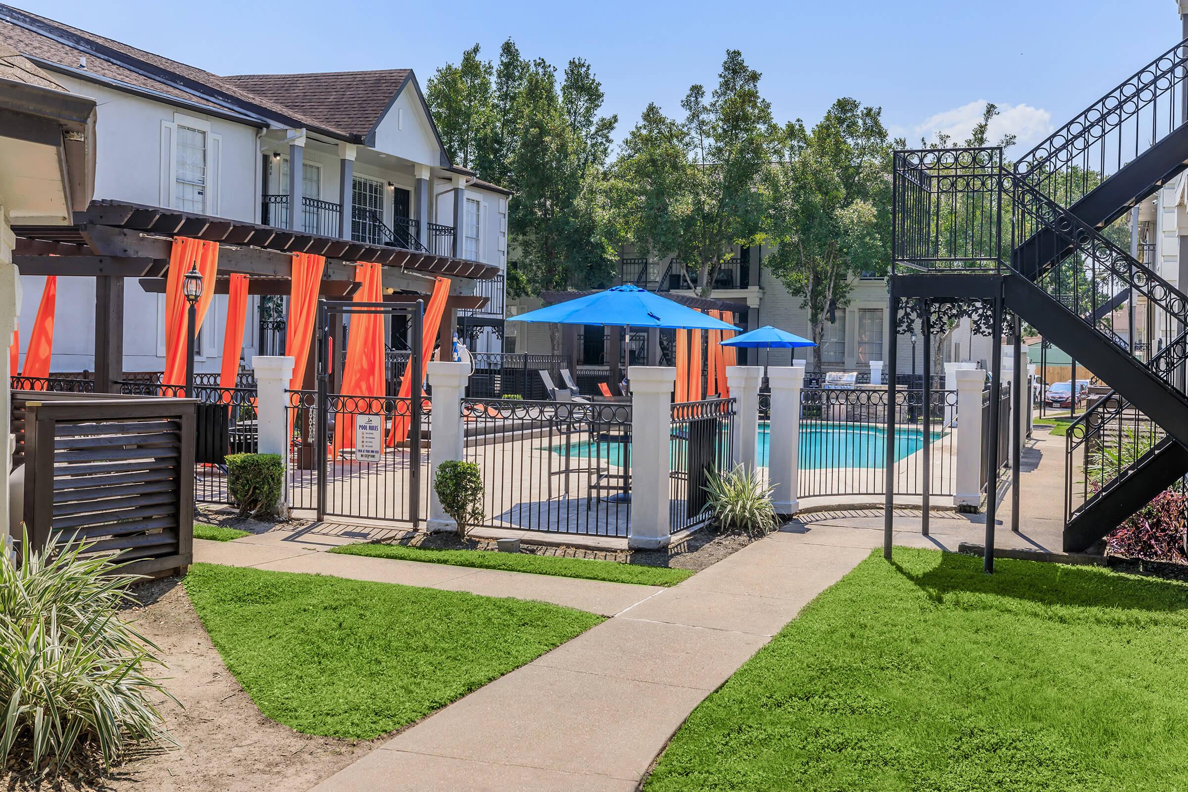 View of a well-maintained outdoor area featuring a swimming pool surrounded by vibrant blue umbrellas. Decorative orange fabric hangs from structures nearby. Lush green grass and pathways lead through the space, with modern architecture visible in the background. A staircase and lounge chairs add to the inviting atmosphere.