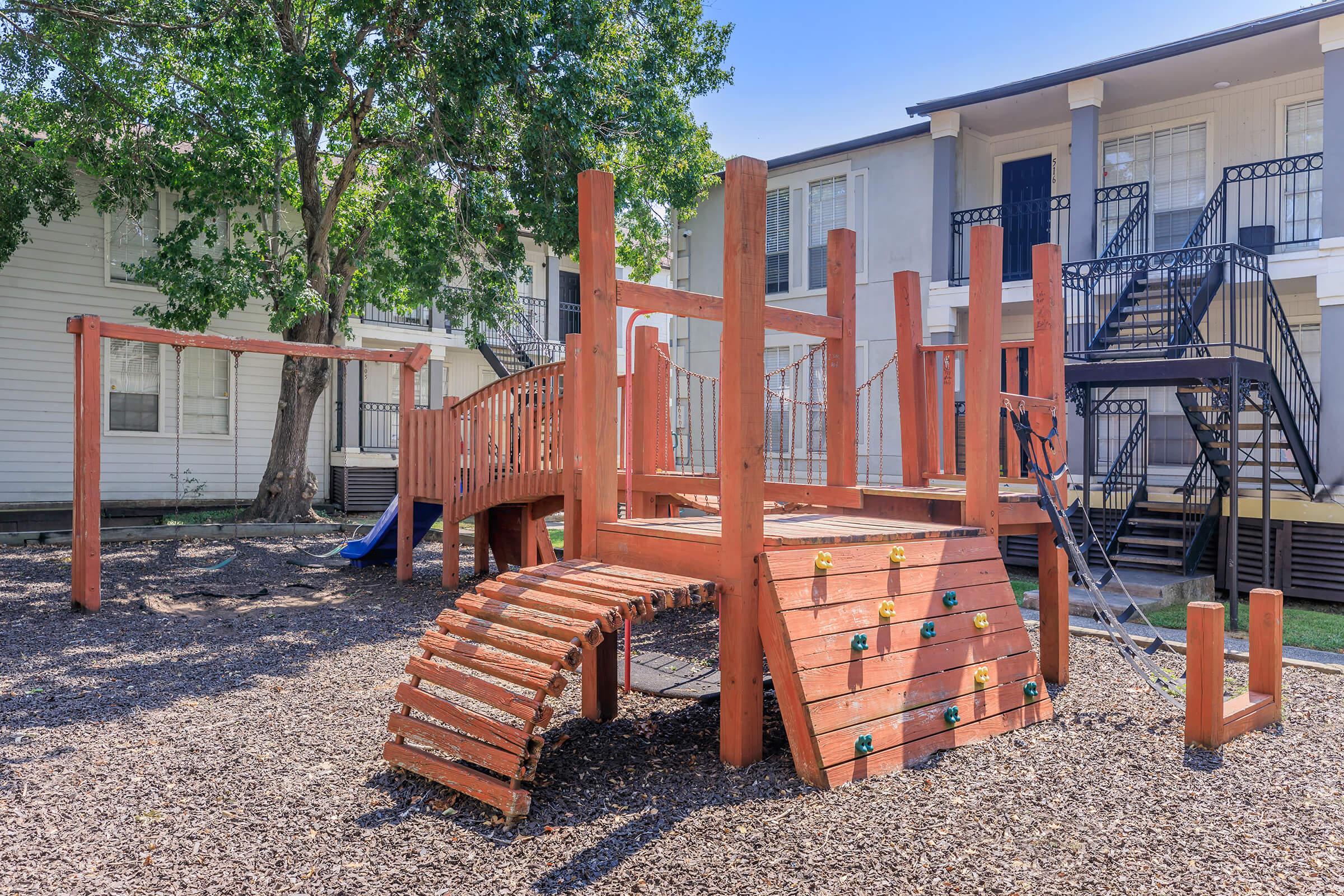 A wooden playground structure with climbing features, slides, and swings situated in a shaded area with mulch. Background includes apartments with balconies and stairs, creating a family-friendly environment suitable for children to play.