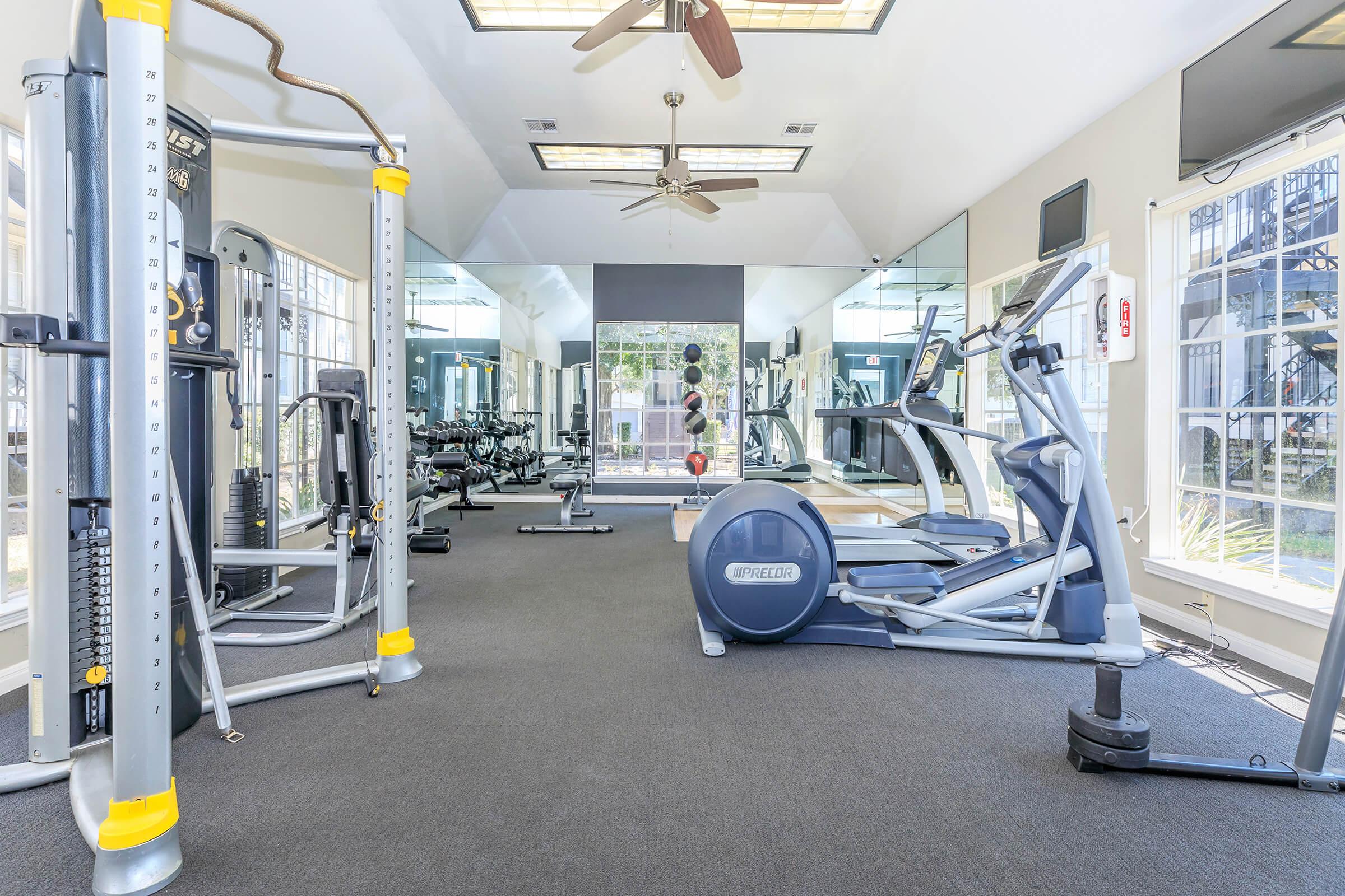 A bright and spacious fitness center featuring various gym equipment such as weight machines, free weights, and cardio machines. Large mirrors reflect the workout area, with ceiling fans providing ventilation. Natural light enhances the atmosphere through windows along the walls.